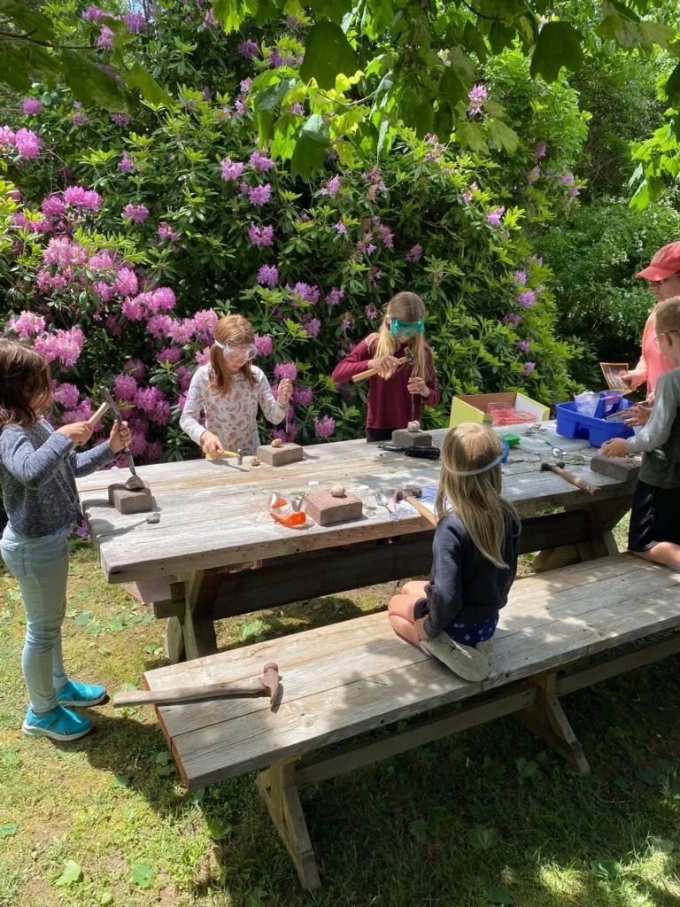 Children engaging in outdoor rock painting activity at a wooden picnic table surrounded by lush green trees and purple flowering bushes.