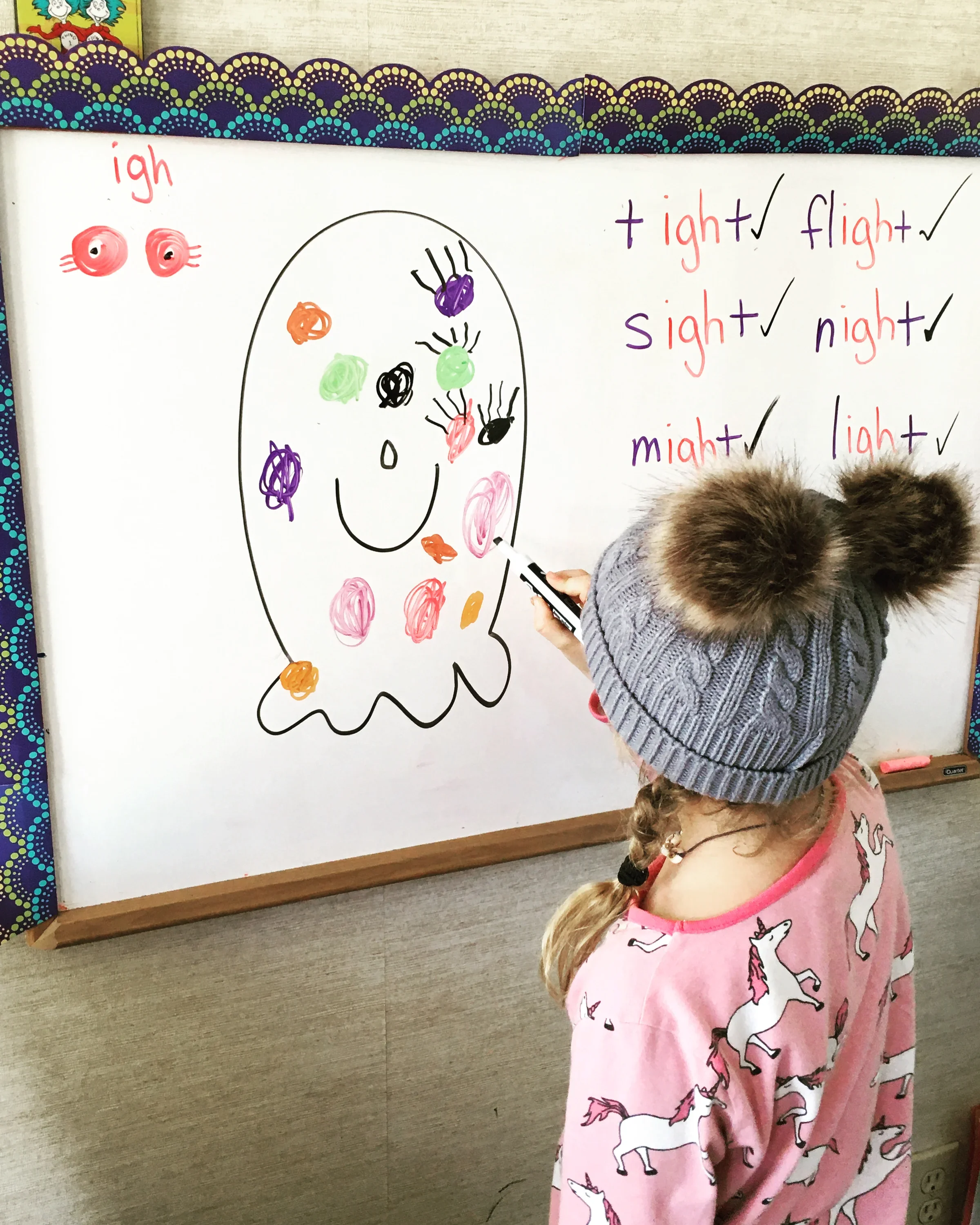 A young girl in a pink pajamas with a unicorn pattern stands in front of a whiteboard, which features a hand-drawn smiley face and colorful scribbles inside the face outline. To the right of the face, there are handwritten words 'light,' 'night,' and 'flight' with check marks, and the words 'sight' and 'might' also with check marks.