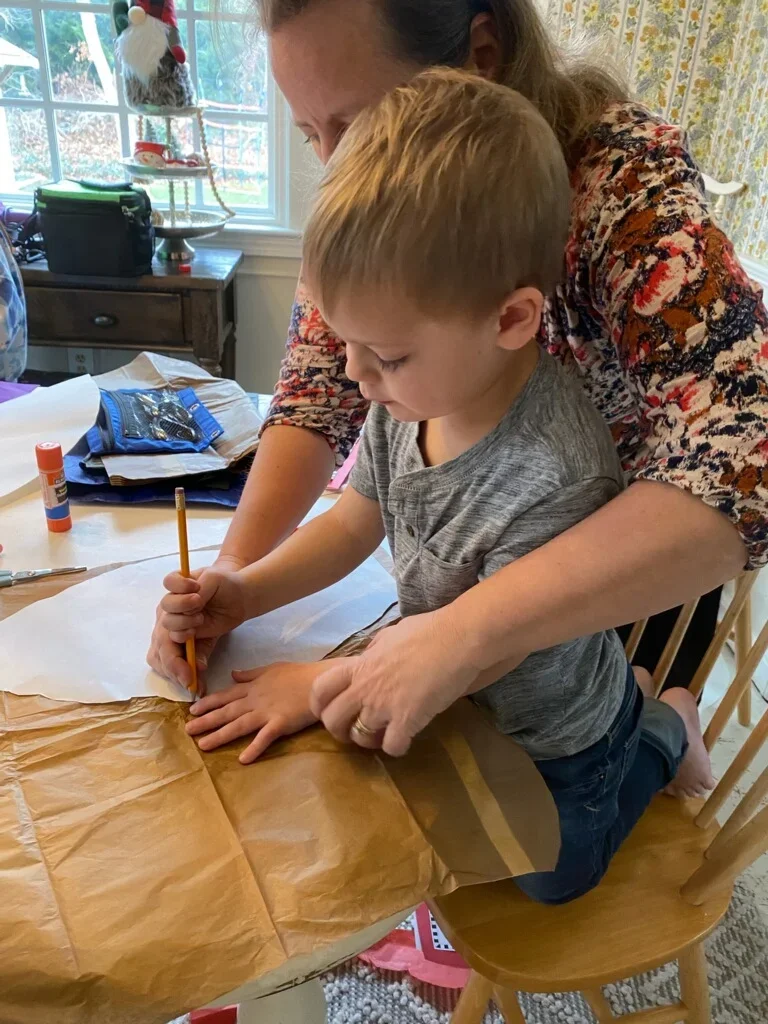 A young boy and an adult woman are at a wooden table, with the woman helping the boy trace on brown craft paper using a pencil. There are craft supplies like glue and scissors on the table, and a window in the background with a wintery outdoor scene.