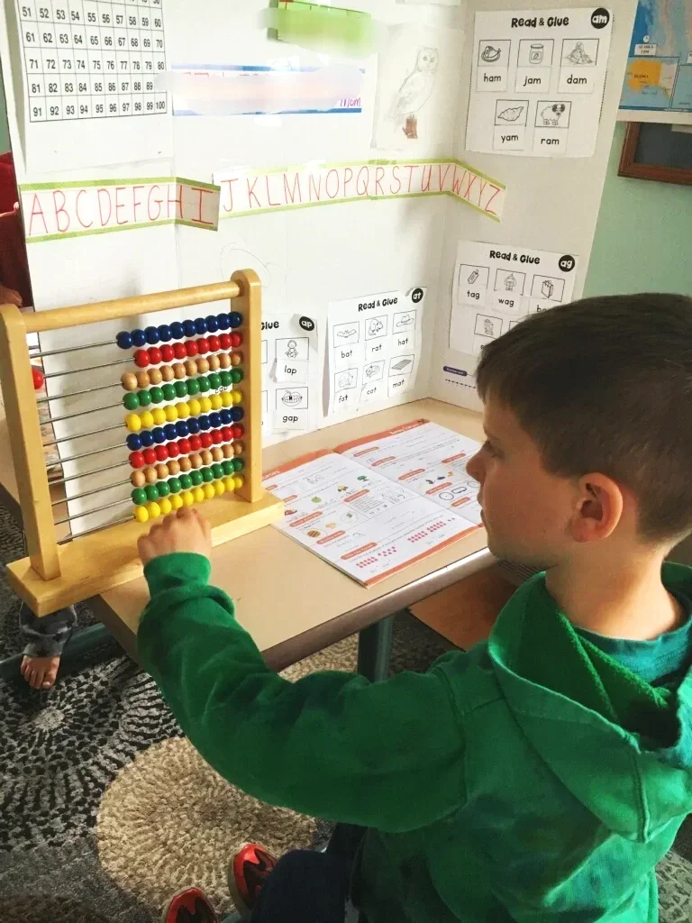 Young boy in green hoodie at classroom desk, using an abacus with colorful beads, educational posters and reading materials on the wall.