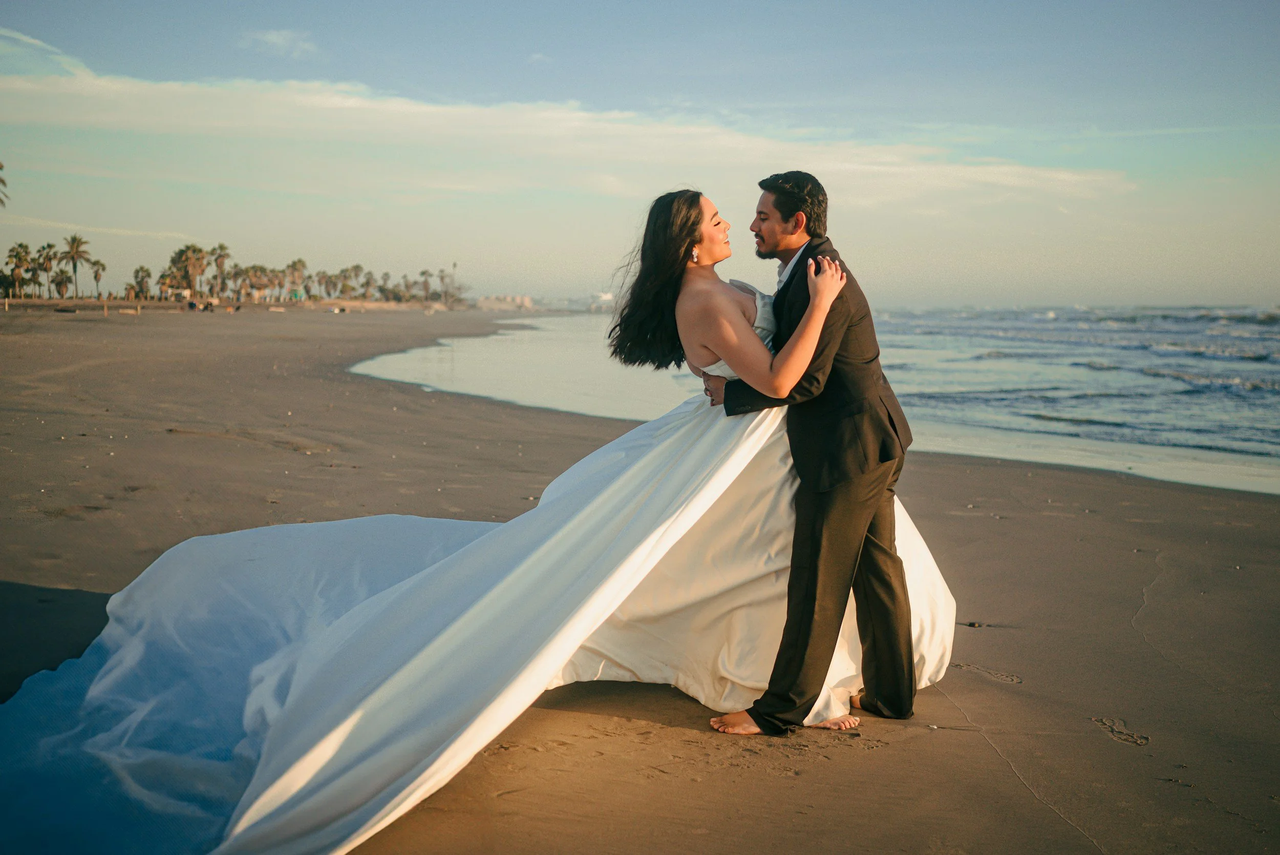 A bride and groom embracing on a beach at sunset, with palm trees and ocean in the background. Beach wedding, Venice Florida, Siesta Key, Nokomis, Englewood, Sarasota