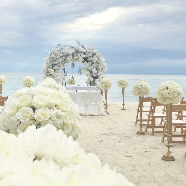 Beach wedding flloral arrangements, wooden chairs, and a floral arch over the aisle on the sand, overlooking the ocean under cloudy skies.