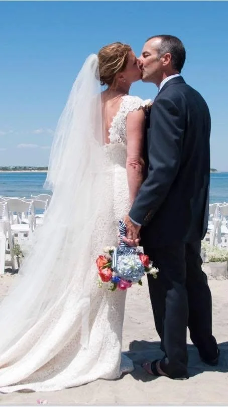 A bride and groom sharing a kiss on the beach after their wedding, with white chairs and the ocean in the background. Beach wedding, Summer wedding.