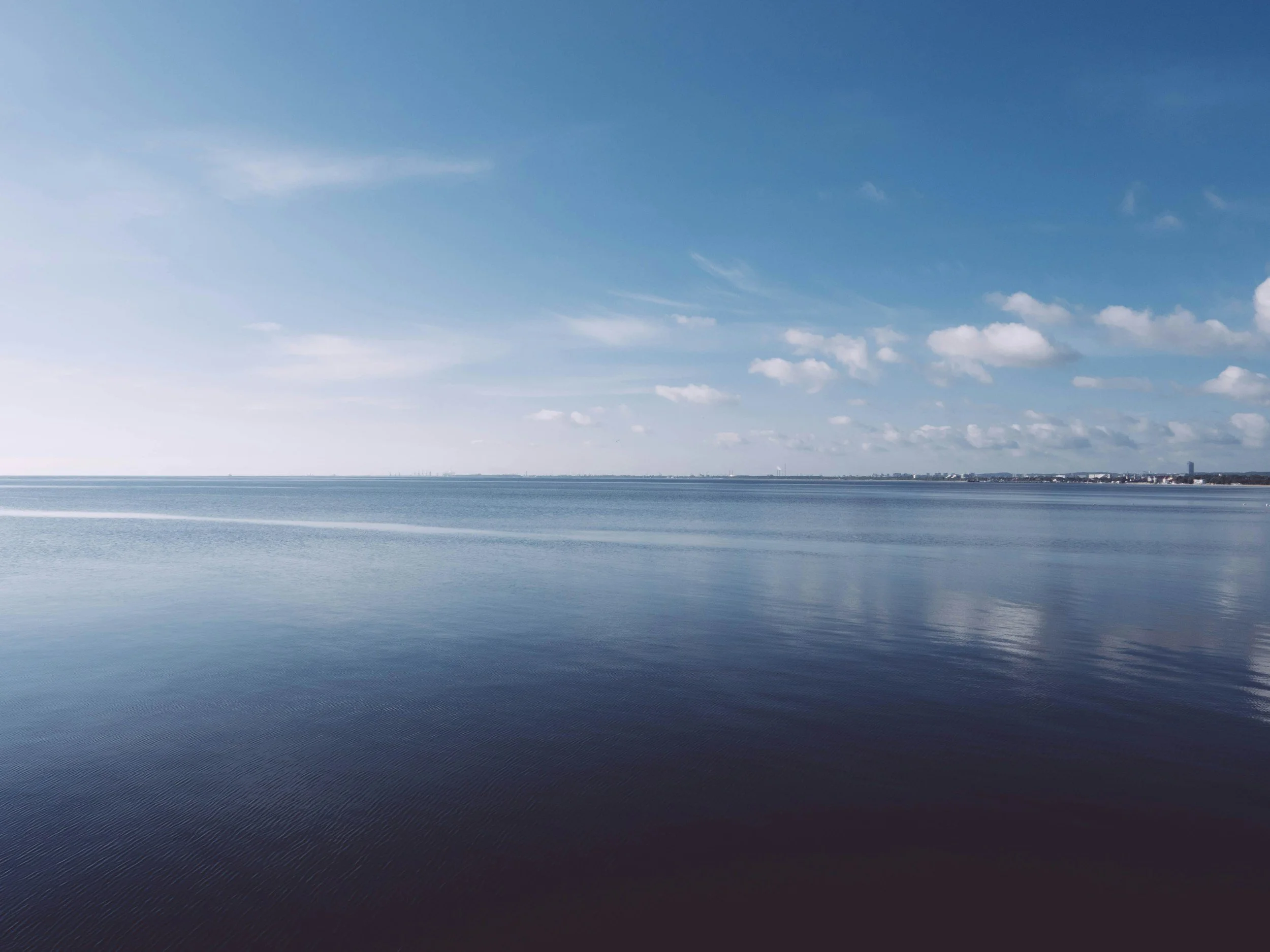 Calm ocean or large body of water under a blue sky with scattered clouds, distant shoreline with buildings and trees.