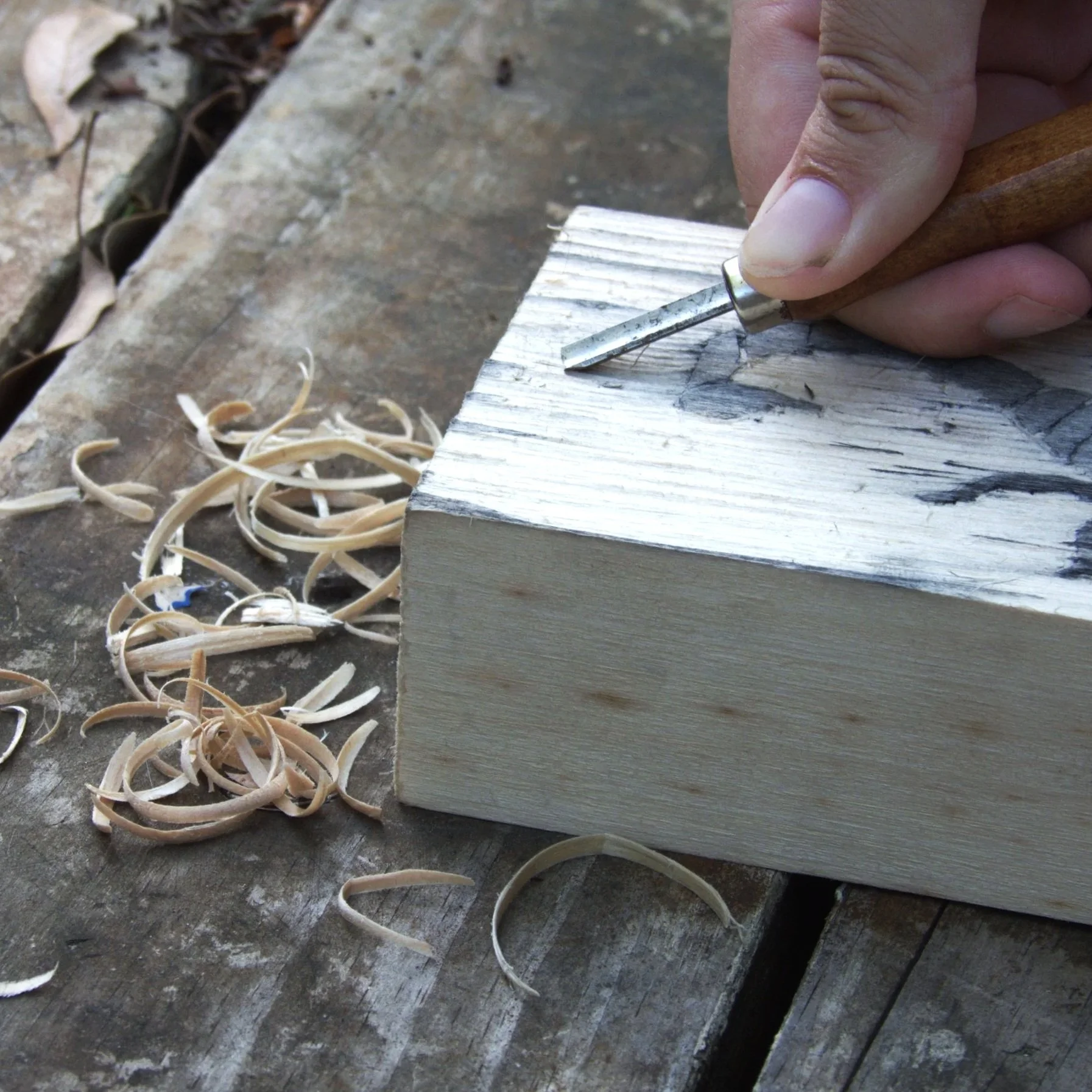 Close-up of a person carving into a piece of wood with a chisel and wooden handle, with wood shavings scattered nearby on a weathered wooden surface.
