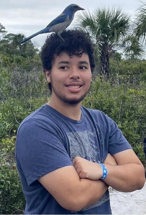 A young man with curly hair standing outdoors with a blue bird perched on his head, greenery and palm trees in the background.