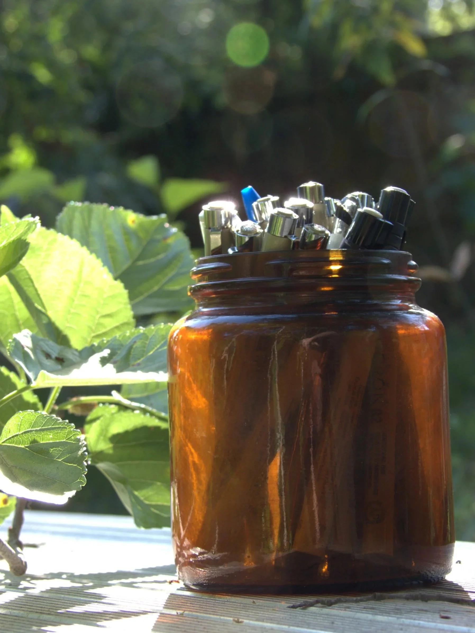 An amber glass jar filled with assorted pens and markers, placed on a wooden surface outdoors with green leaves and sunlight in the background.