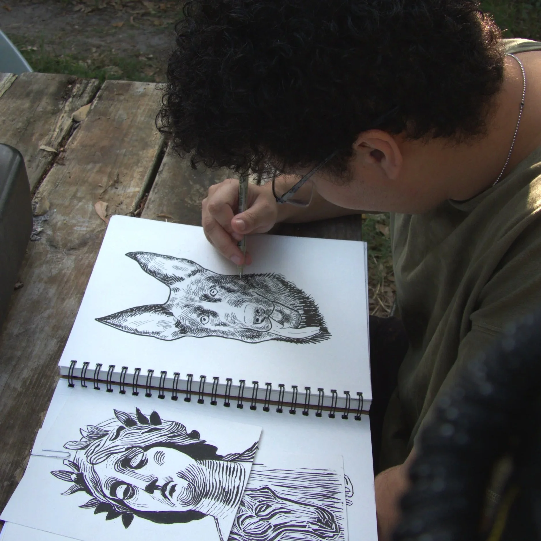 An artist sketching a detailed black and white drawing of a dog's face in a sketchbook on a wooden table outdoors.
