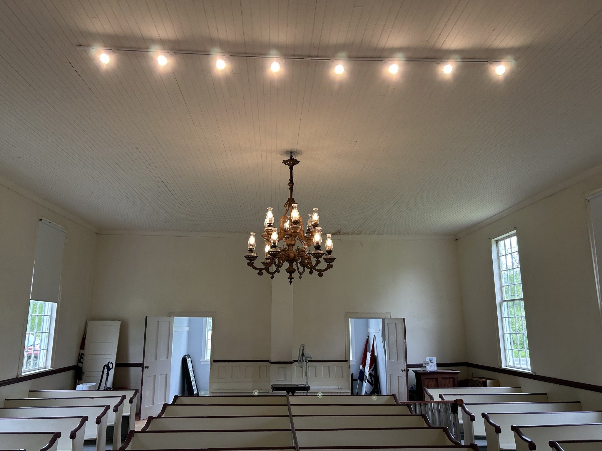 Empty church with white pews, a chandelier hanging from a wooden paneled ceiling, and windows on the side walls.