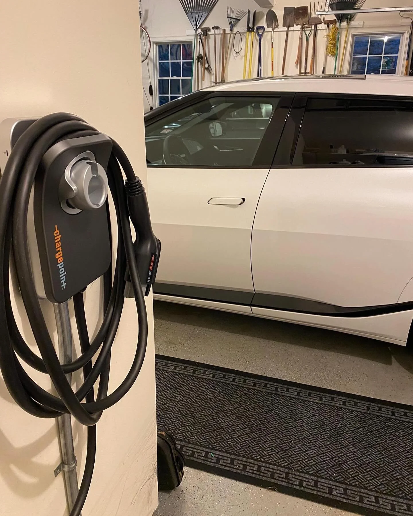 Electric vehicle charging station with a charging cable and plug in a garage, with a partial view of a white car and garage tools on the wall.