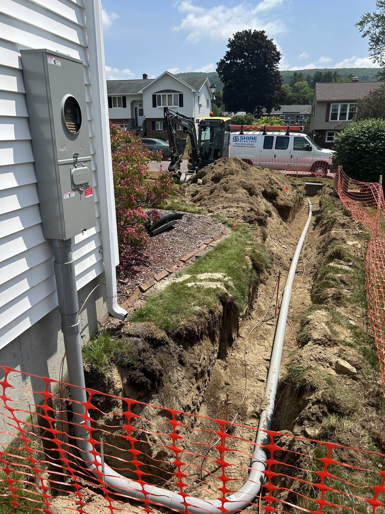 A residential construction site with a trench for underground piping. A pipe runs through the trench, which is surrounded by orange safety mesh. A small excavator and a white utility van are in the background, with houses and a hill visible under a p
