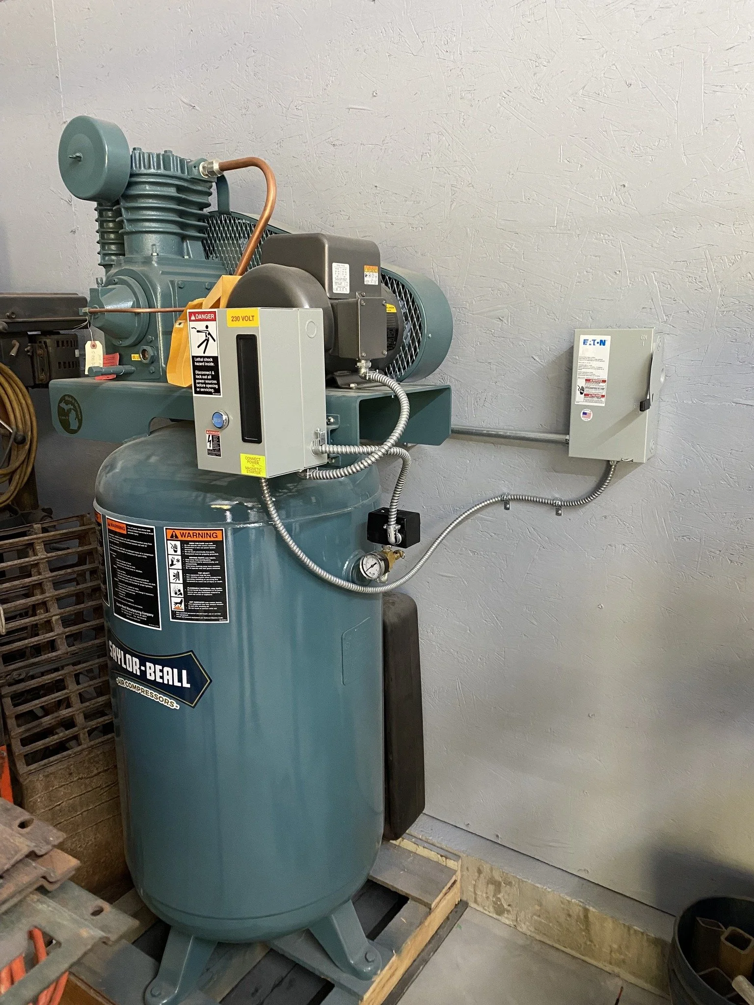A large turquoise air compressor with various labels and gauges, positioned on a wooden pallet against a beige wall.