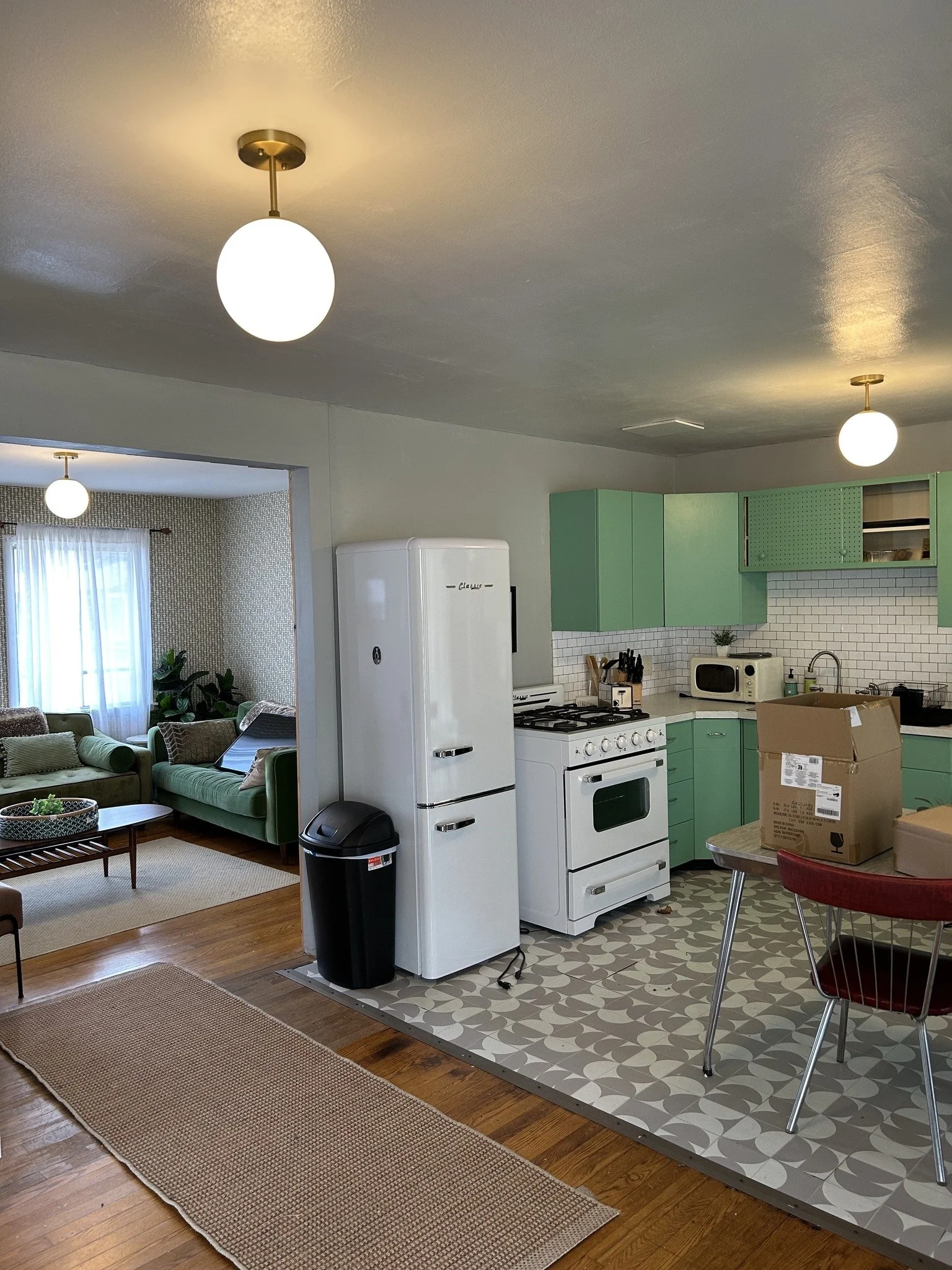 View of a kitchen with green cabinets, white appliances, and a patterned tile floor. Adjacent living area with sofas and large window. New lighting.