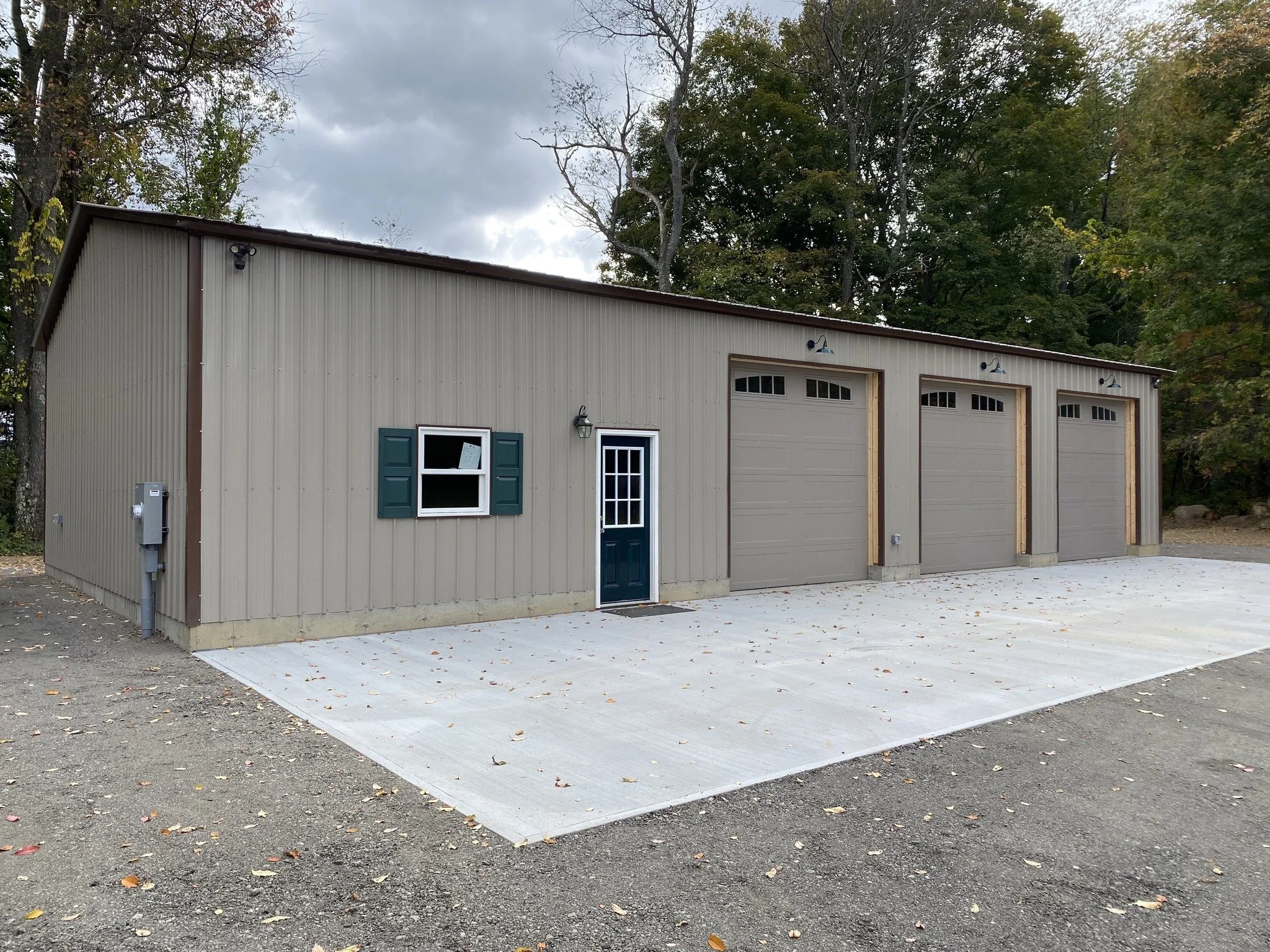 A beige metal garage building with three garage doors, a small green door, a window with green shutters, and outdoor lighting, surrounded by trees with autumn foliage.