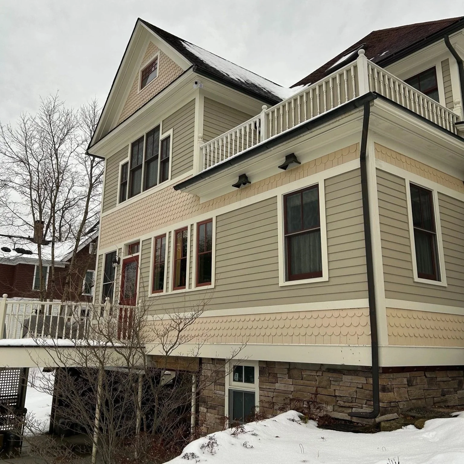Side view of a multi-story house with beige siding, stone foundation, snow on roof, and leafless trees.
