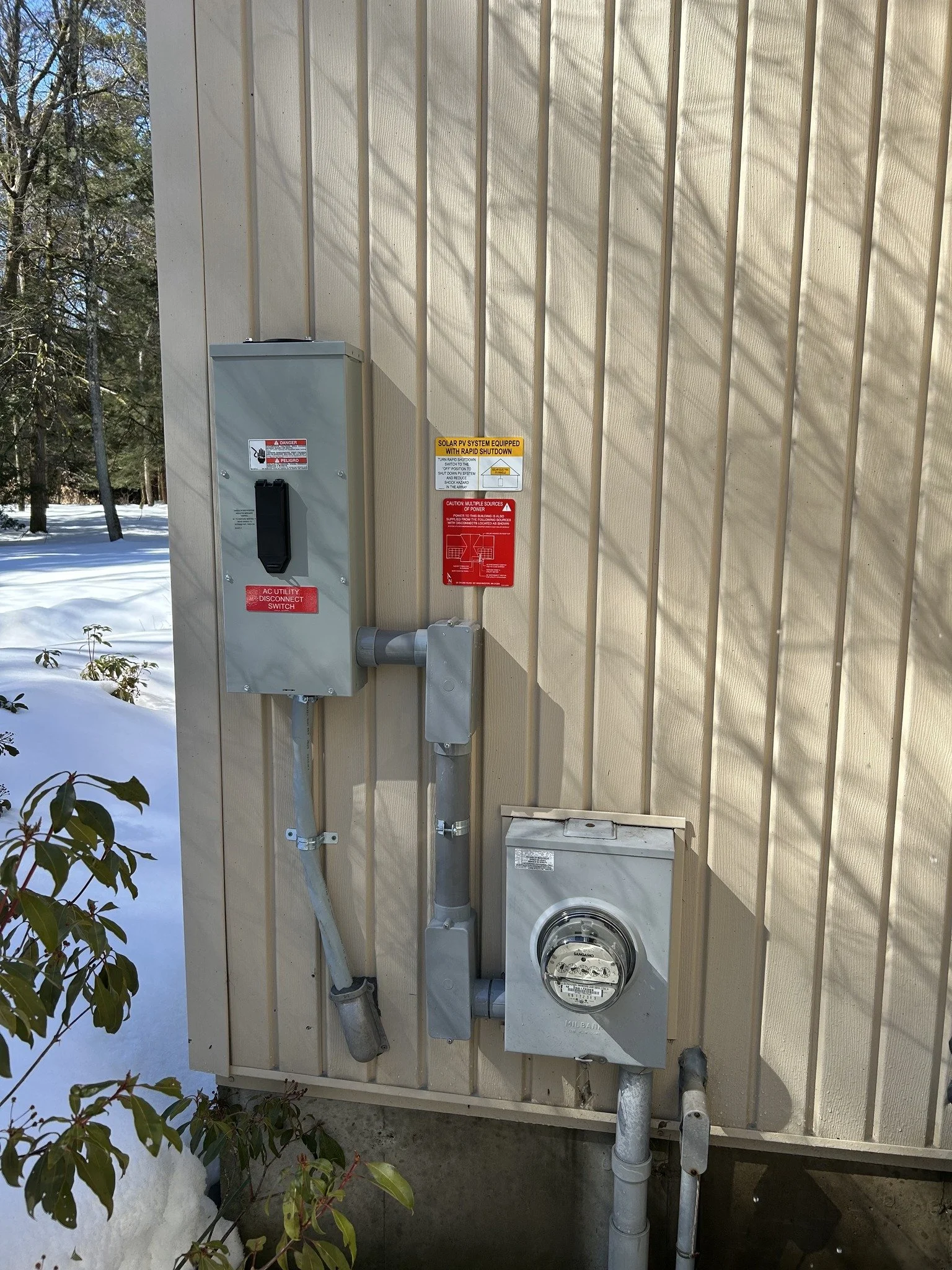 Outdoor electrical and utility boxes attached to the beige siding of a building, with snow on the ground and trees in the background.