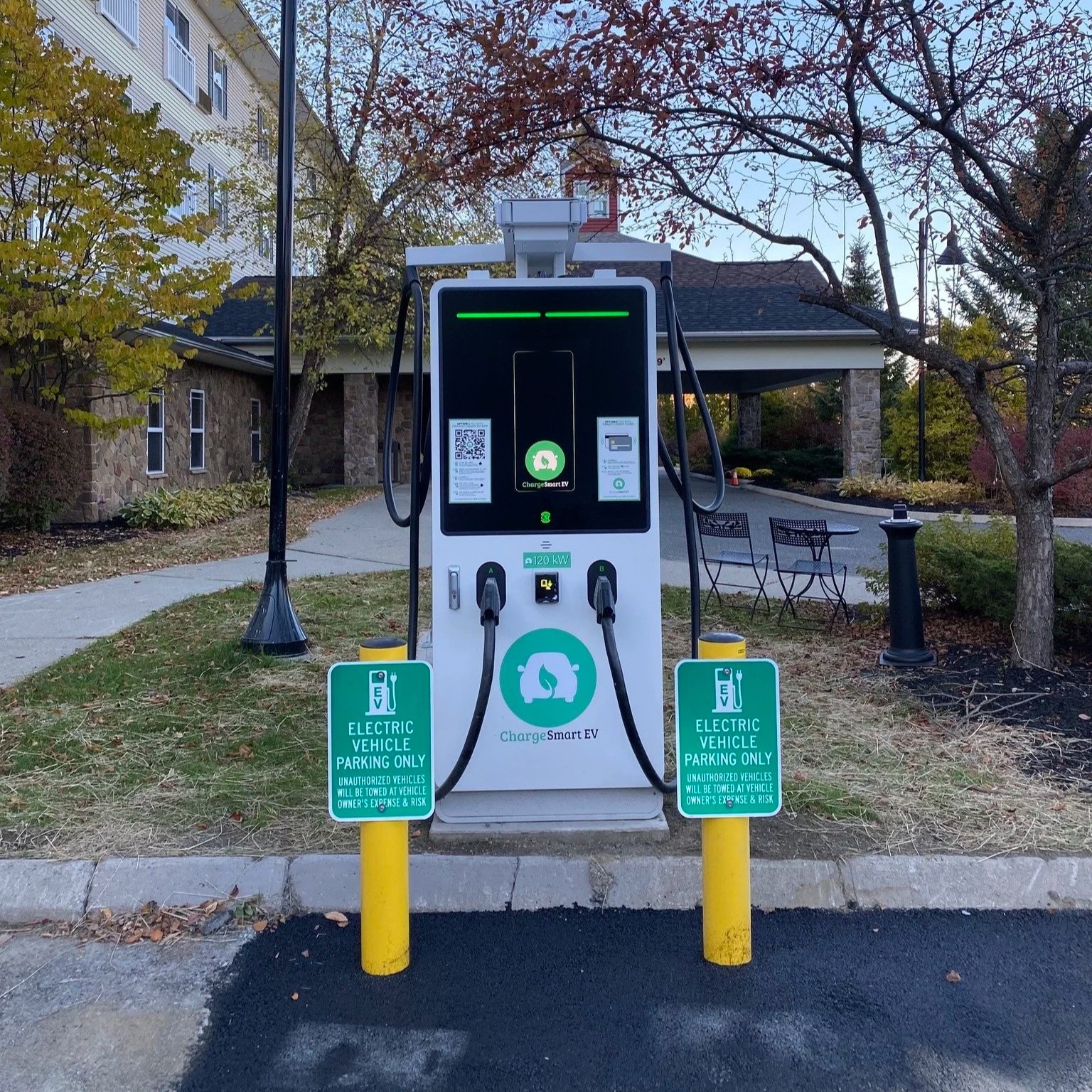 Electric vehicle charging station with two charging cables, surrounded by yellow safety bollards, in an outdoor parking area with trees, benches, and a building in the background.