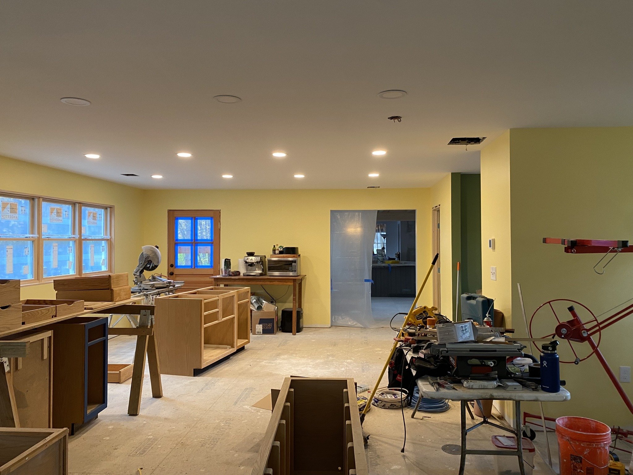 Interior of a room under renovation with yellow walls, unfinished wooden furniture, tools, and construction materials scattered around, including a miter saw, table, and various supplies.