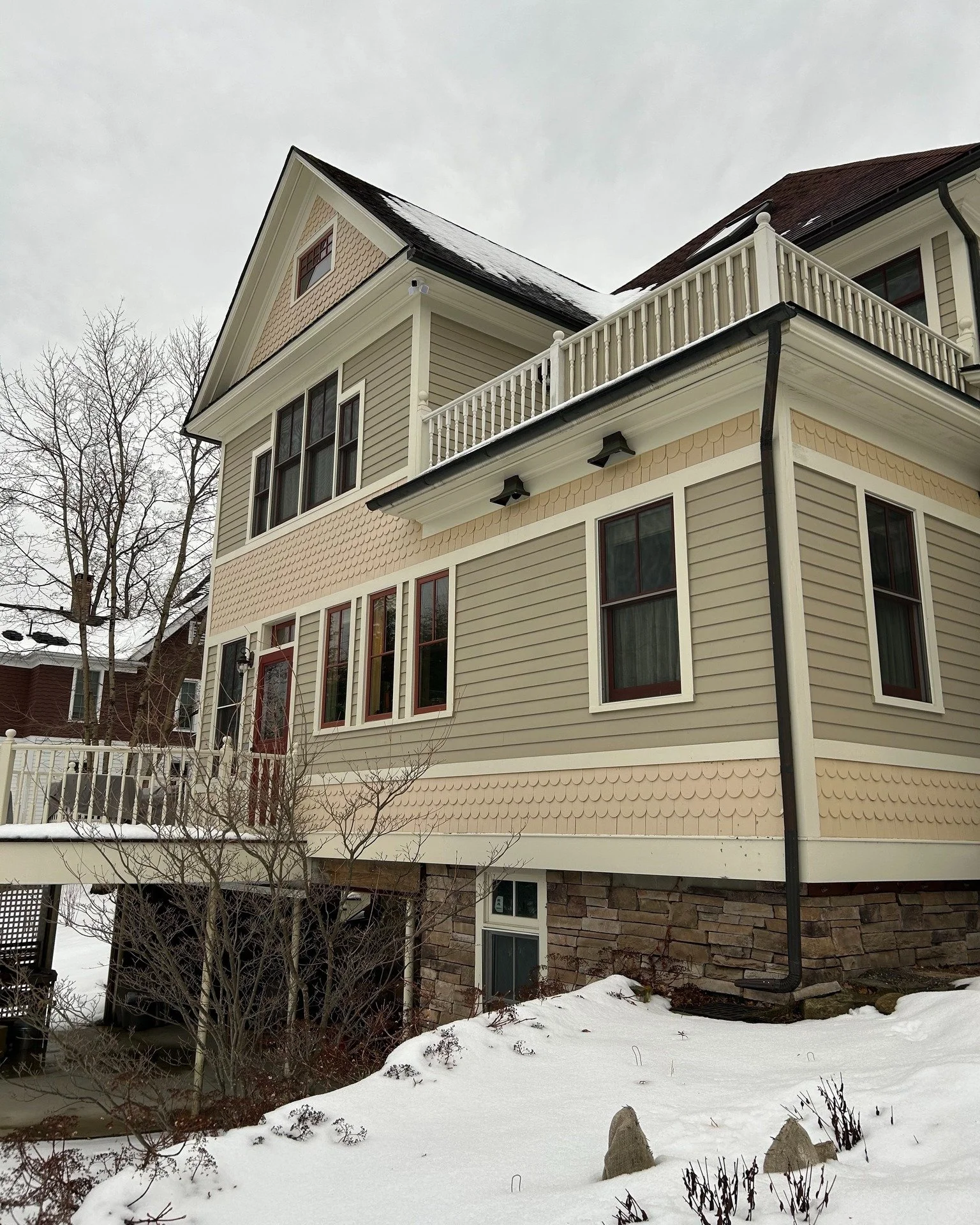 Front view of a multi-story house with beige and cream siding, surrounded by snow, with a stone foundation and leafless trees nearby.