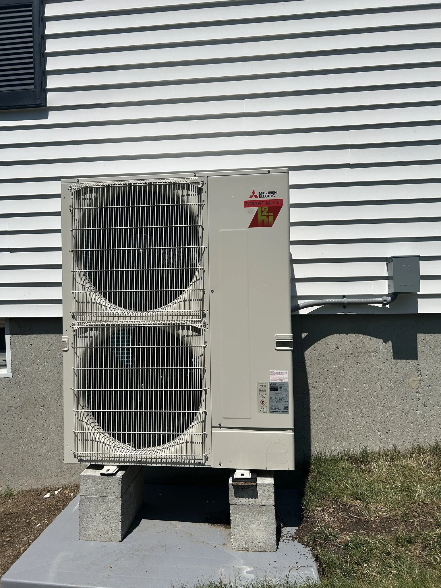 Outdoor Mitsubishi Electric air conditioning or heat pump unit mounted on concrete blocks in front of a white house with horizontal siding.
