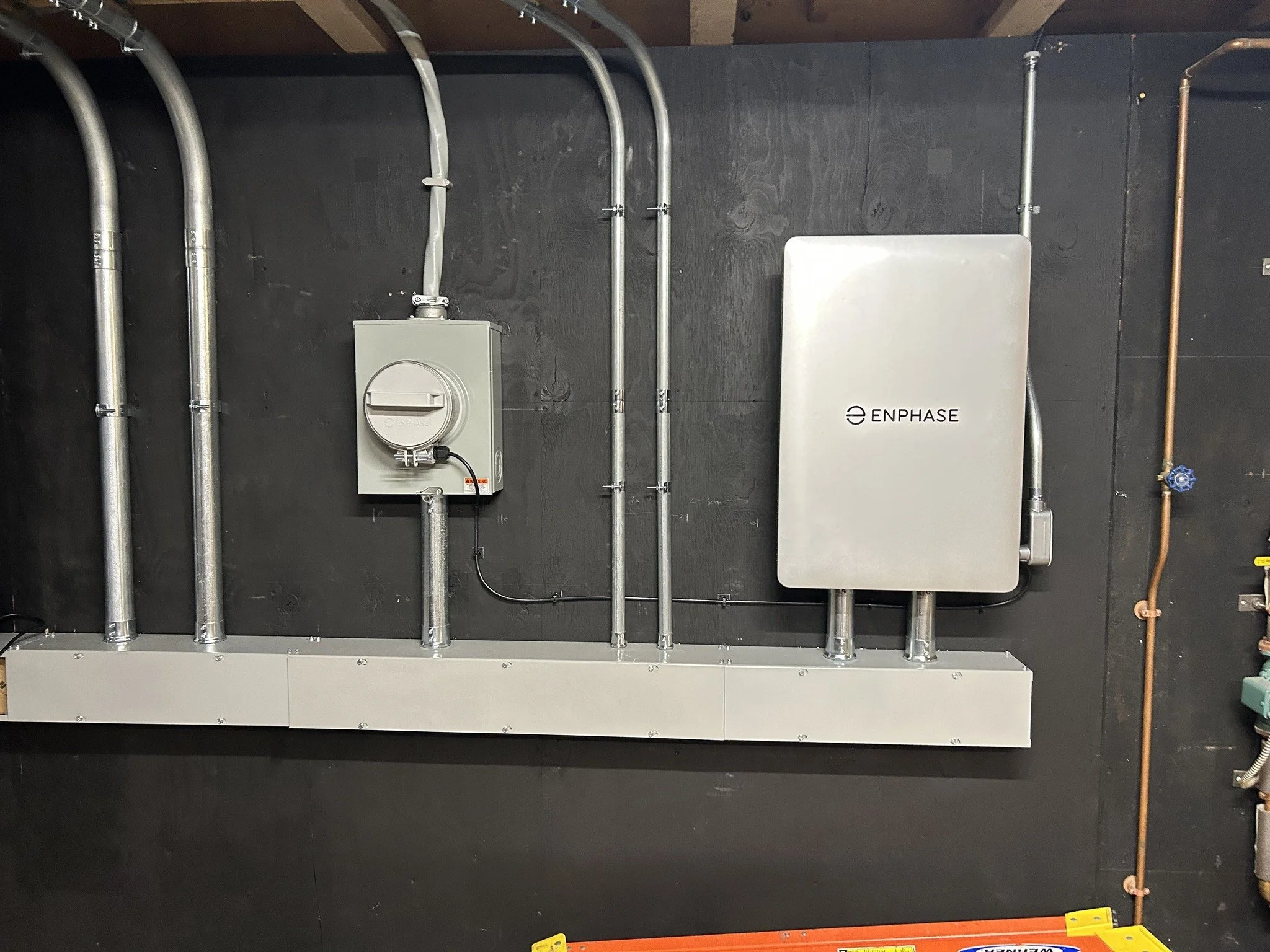 Electrical panel and equipment mounted on a black wall, including a gray box with a dial, metal conduit pipes, and an ENPHASE solar inverter or power control device.