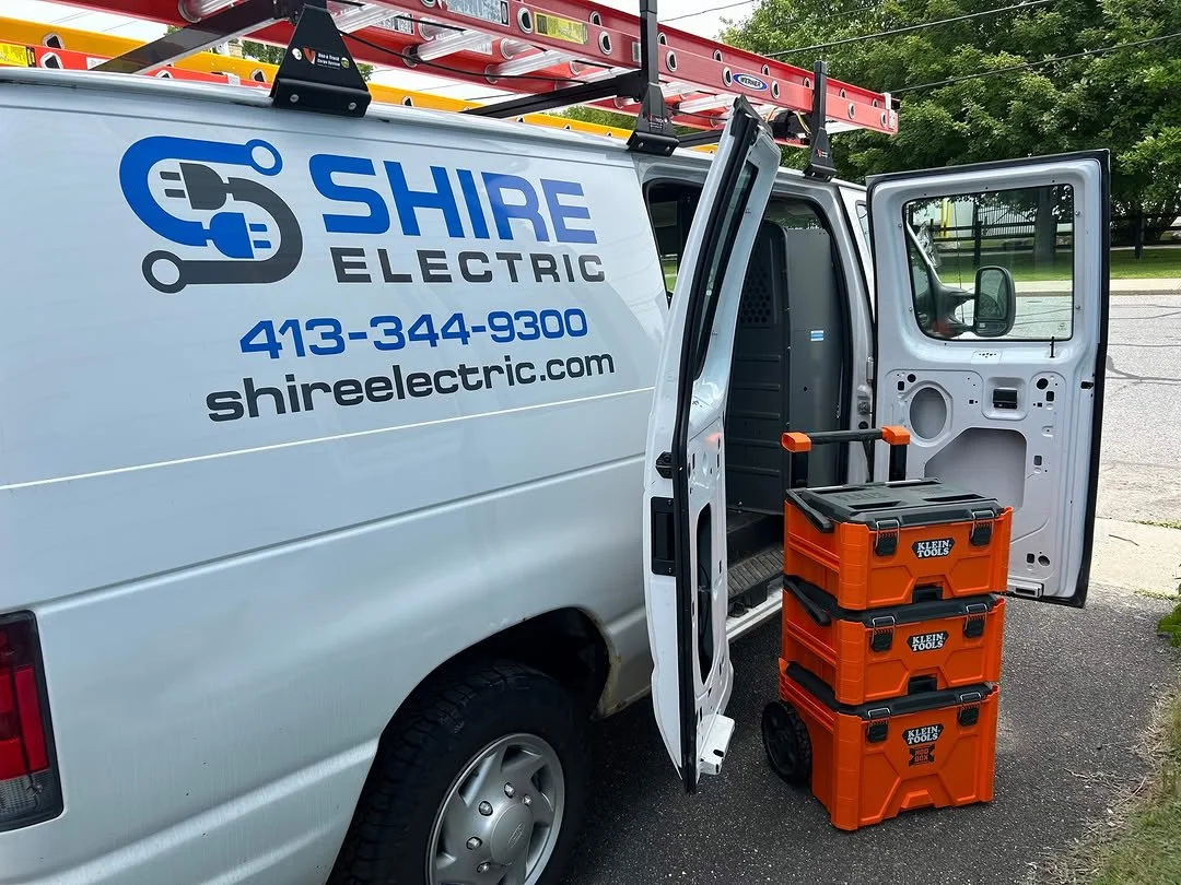 A white van with blue and black lettering for Shire Electric, displaying a phone number and website, parked outdoors next to some orange tool boxes on the ground, with a ladder and trees visible in the background.
