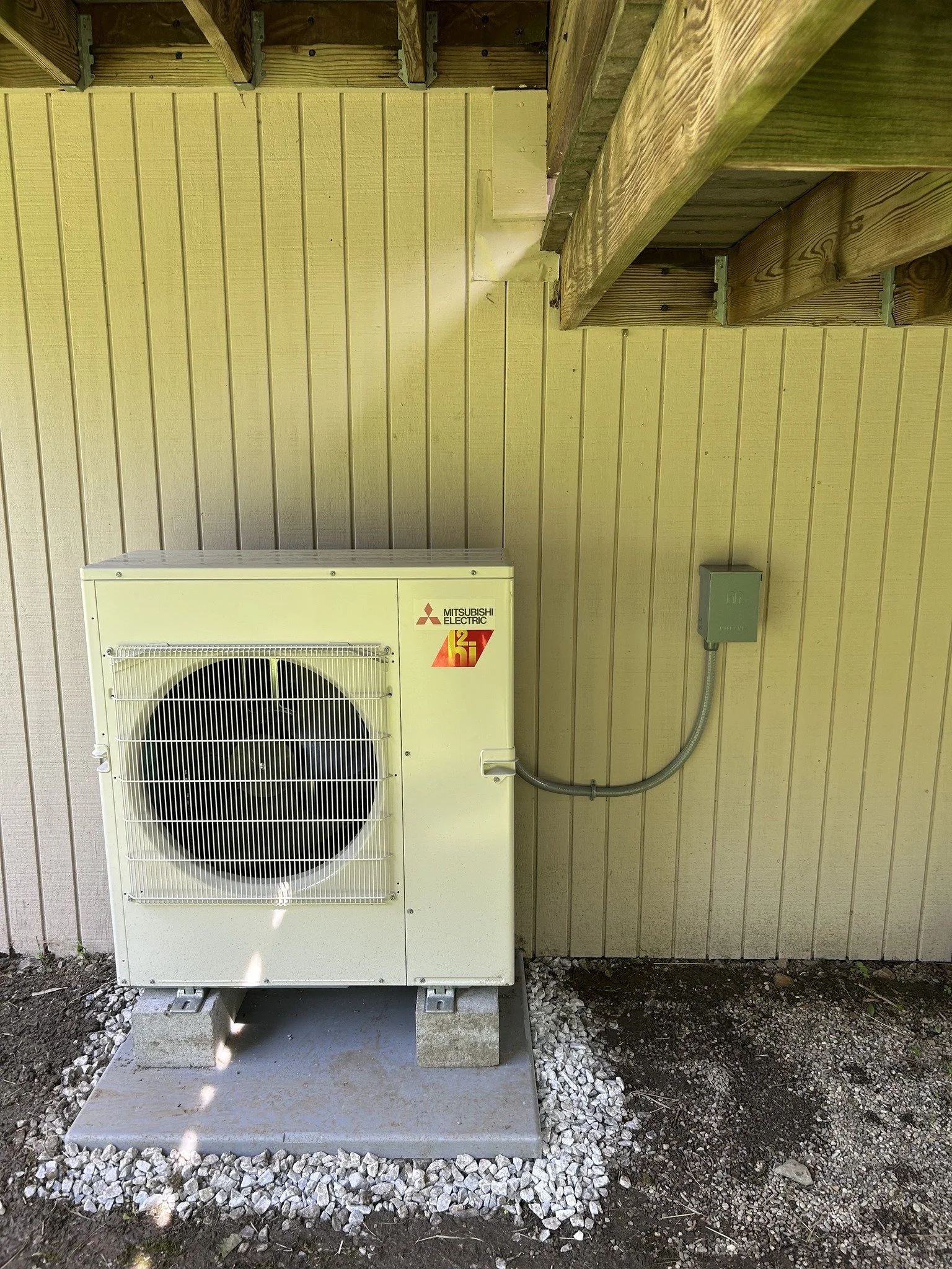 Outdoor Mitsubishi Electric HVAC unit on a concrete pad with gravel, next to a yellow wall and wooden porch above.