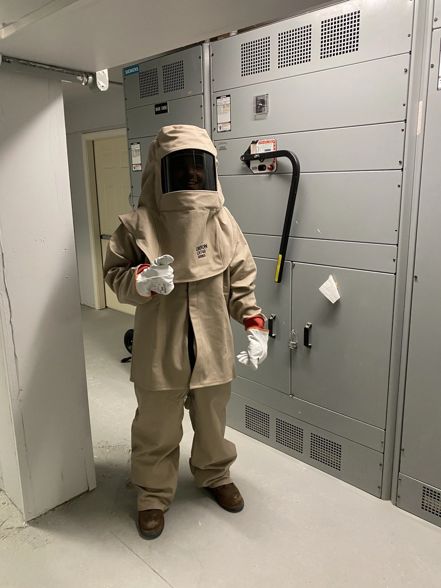 Person wearing a beige hazmat suit, gloves, and face shield standing in front of electrical equipment.