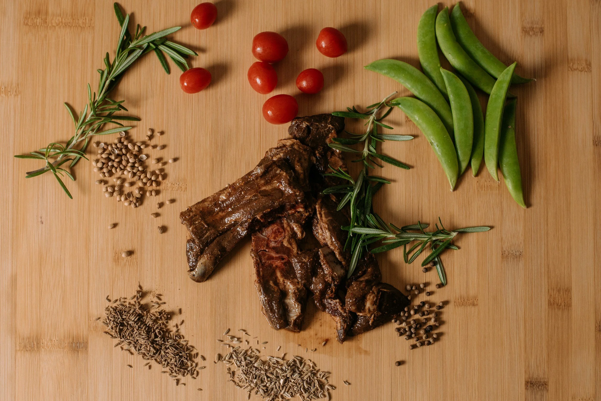 Raw beef ribs surrounded by cherry tomatoes, green beans, rosemary, sage, cumin seeds, and black pepper on a wooden cutting board.