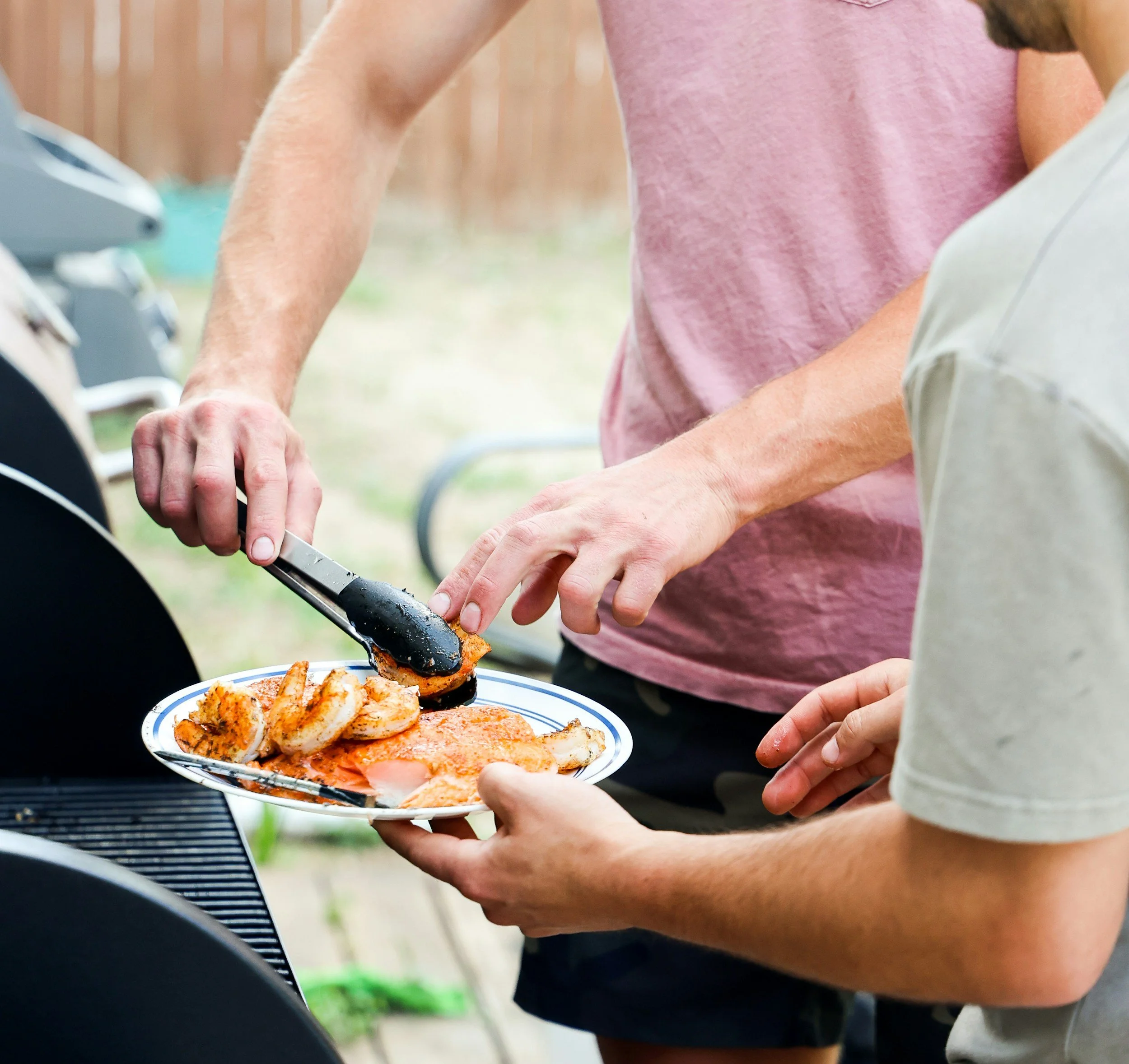 A person is using tongs to place cooked shrimp and salmon on a plate, with a grill in the background.