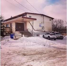 A white building with a sloped roof, surrounded by snow, with a few cars parked in front and a blue trash bin nearby.