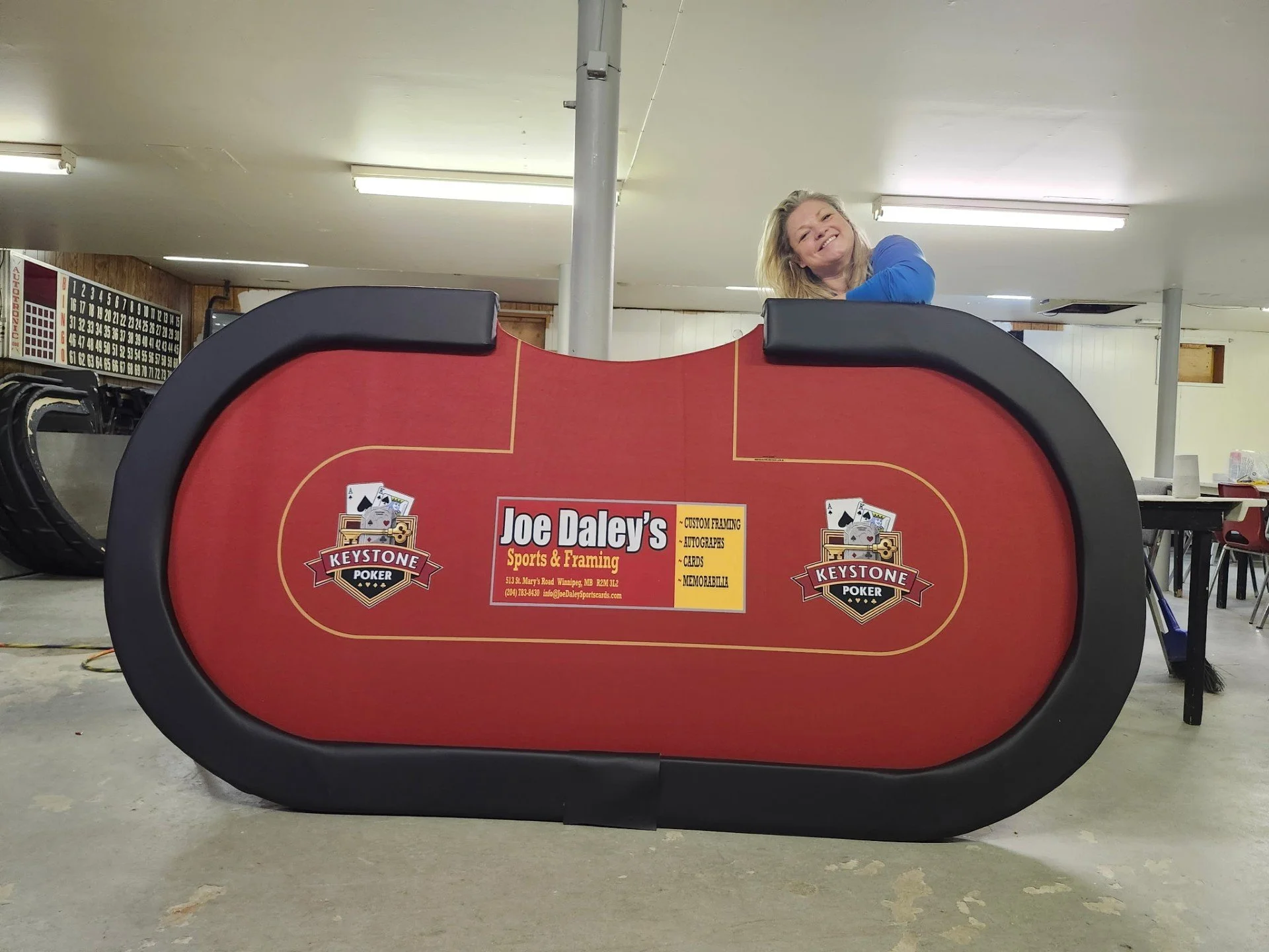 A red poker table with a black border featuring the Joe Daley's Sports & Framing logo, situated in a room with fluorescent lights and wooden-paneled walls. A woman in a blue shirt is smiling and leaning over the table from behind. A bingo game board is visible on the wall to the left.