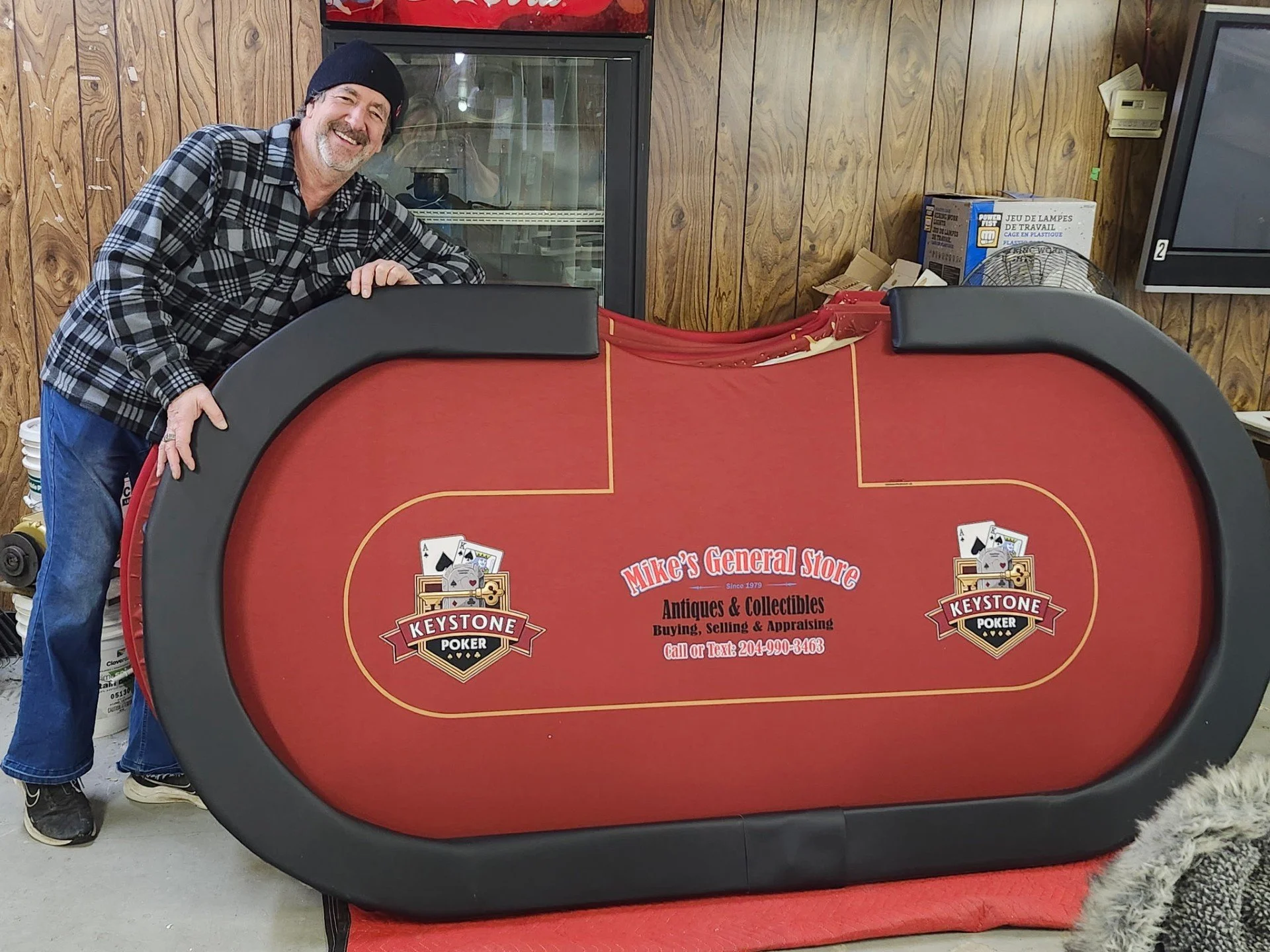 A man wearing a black beanie and a plaid shirt is smiling, leaning on a red poker table with the logo 'Keystone Poker' and the text 'Mike's General Store, Antiques & Collectibles, Buying, Selling & Appraising' at a gaming or entertainment venue with wood-paneled walls.