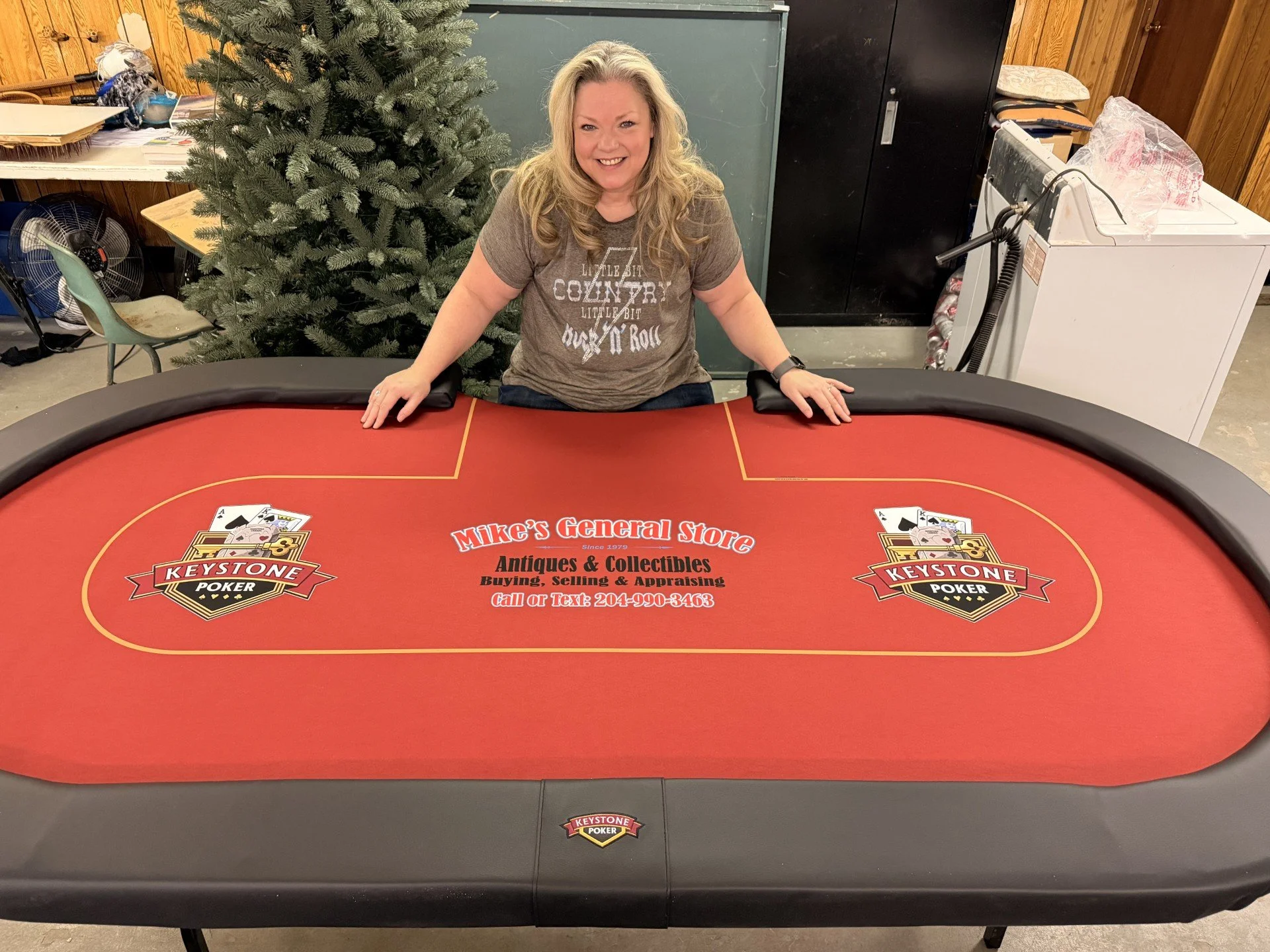 A woman smiling and leaning on a red Keystone Poker table with a black padded railing in a room with wood-paneled walls. There is a Christmas tree to her left, a fan and chair to her right, and various objects including a washer and storage cabinet i