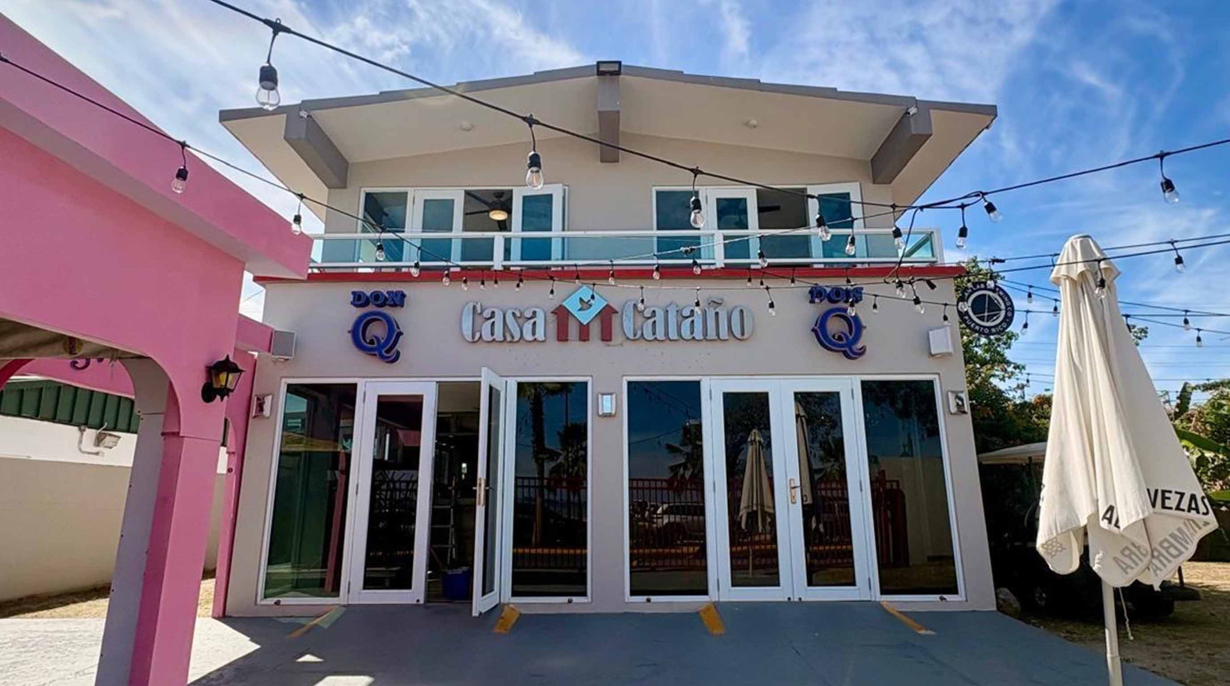 Exterior view of Casa Catalino restaurant with string lights overhead, pink and white walls, and open glass doors.