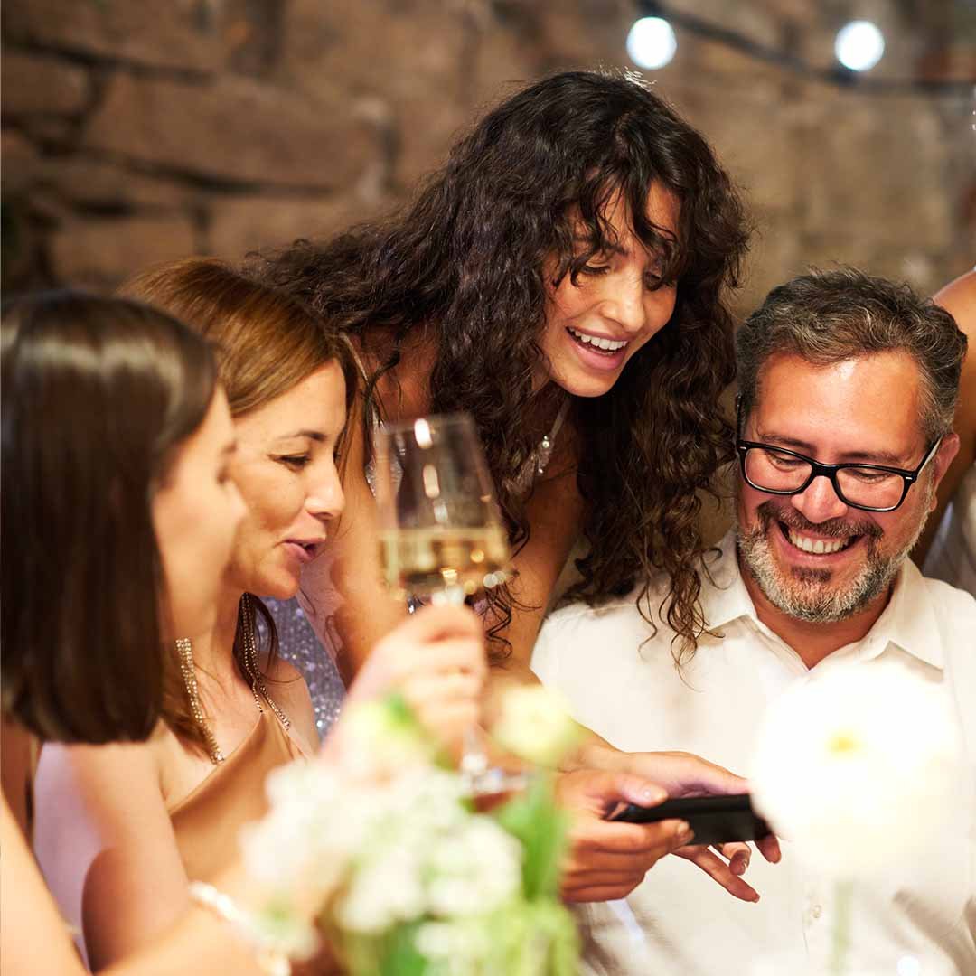 Group of people enjoying a social gathering, looking at a smartphone with smiles, one woman holding a glass of wine.
