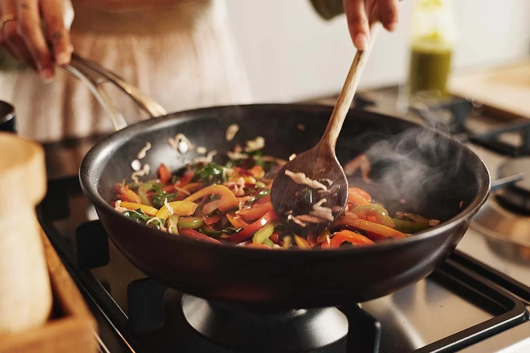 Someone cooking a stir-fry with mixed vegetables in a black frying pan on a stove.