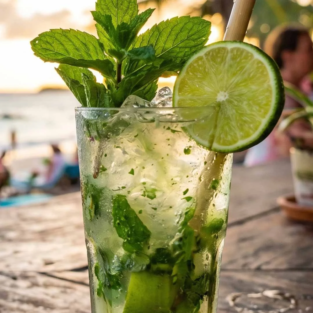 A glass of mojito cocktail with lime wedge and mint leaves, outdoors near the water during sunset.