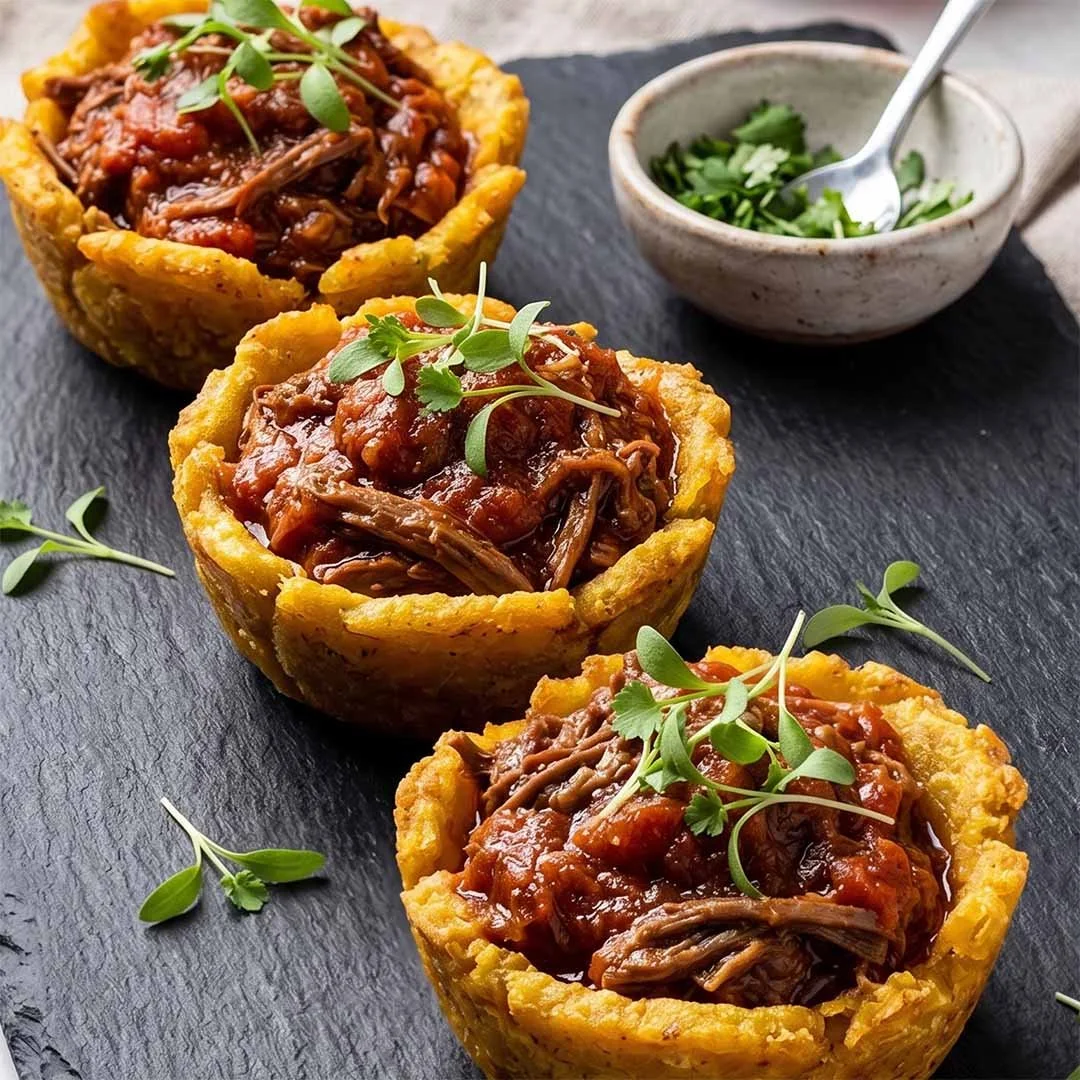 Three cornbread cups filled with shredded beef stew topped with microgreens, served on a black slate board with a small bowl of chopped green herbs in the background.