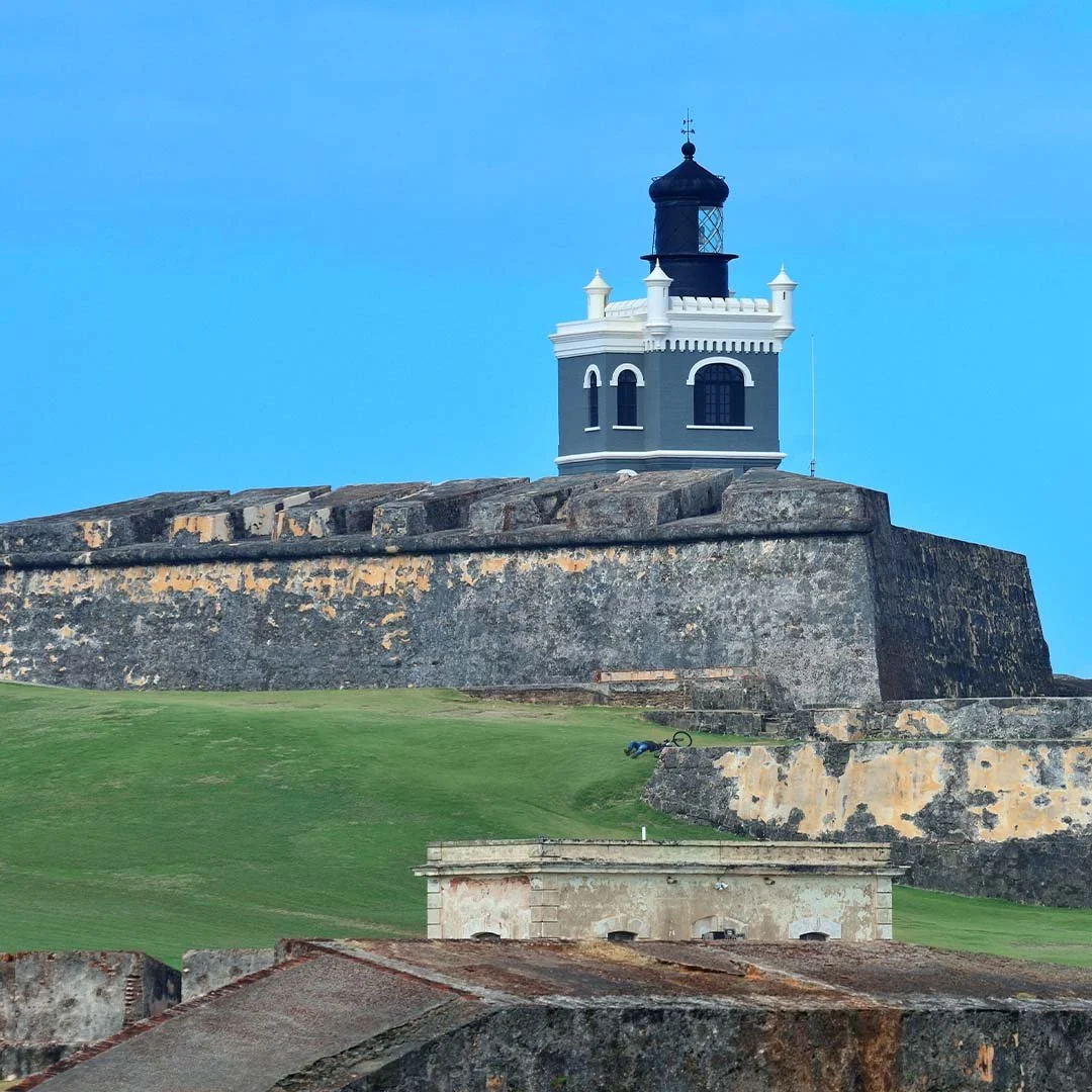 A lighthouse on top of a stone fortress with a green grassy hill in the foreground and a clear blue sky.