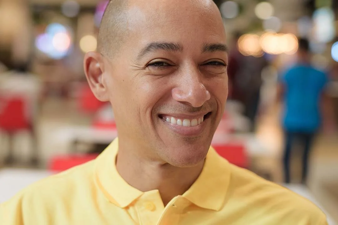 A smiling man with a shaved head wearing a yellow polo shirt, in an indoor setting with blurred lights and people in the background.