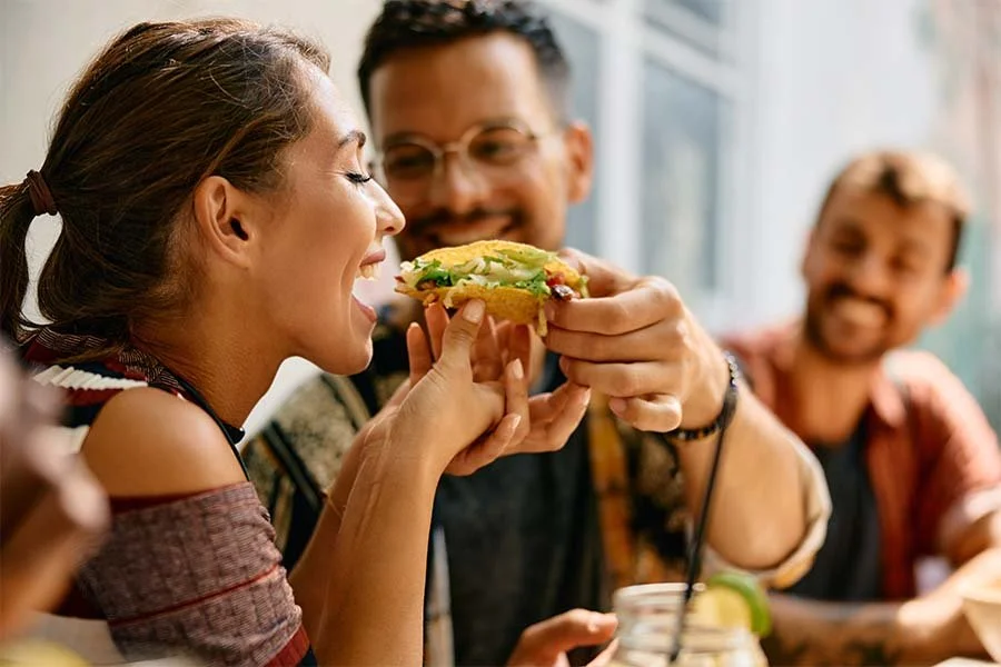 A woman about to bite into a sandwich held by a man, with two other smiling people in the background at a casual gathering.