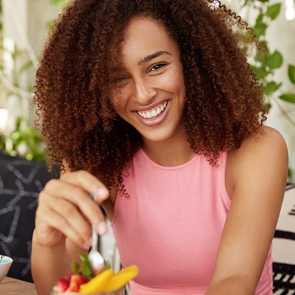 A young woman with curly hair smiling, wearing a pink sleeveless top, sitting at a table with a bowl of fruit, in a bright, plant-filled room.
