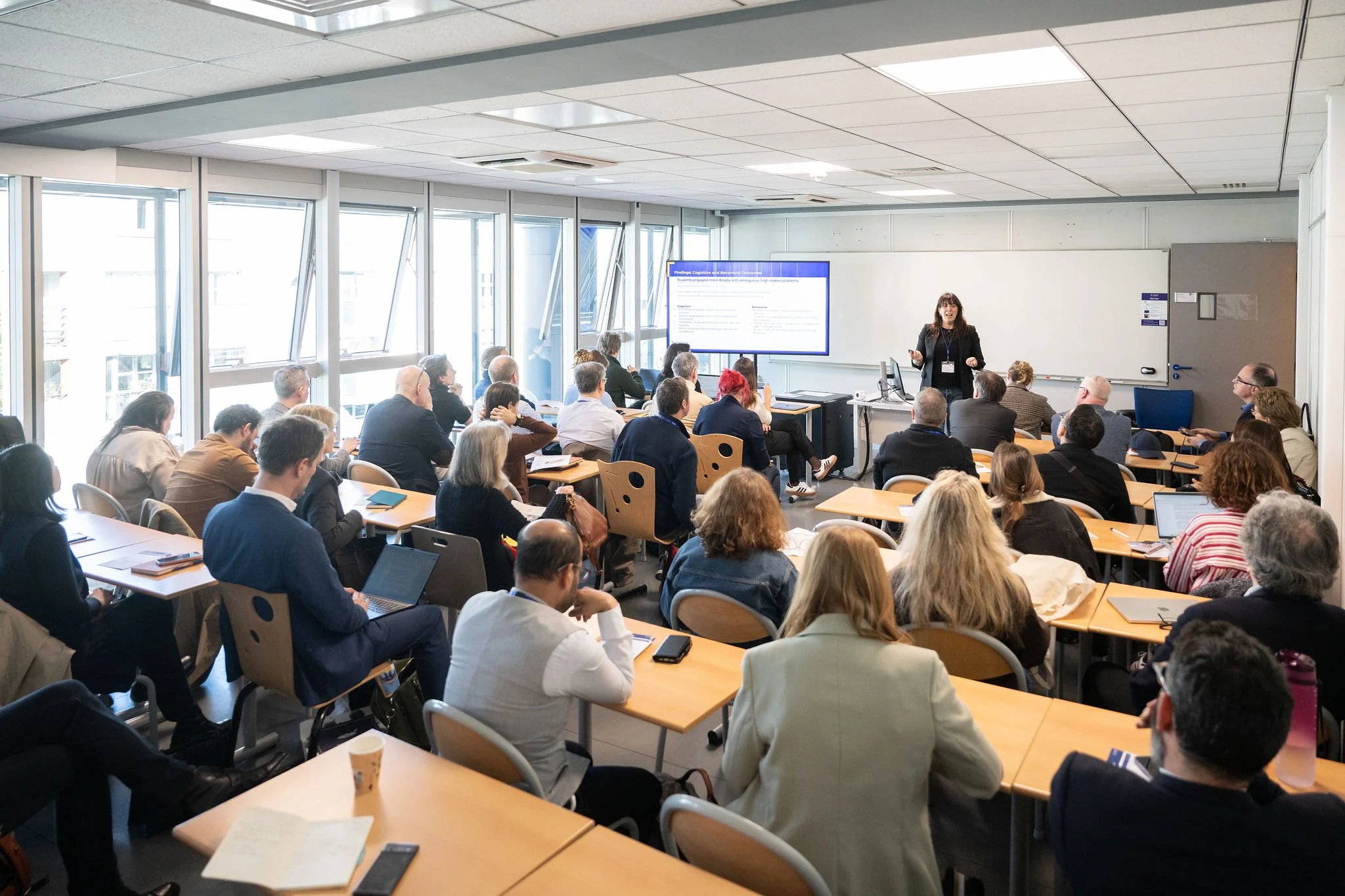 A diverse group of people attending a professional presentation or conference in a well-lit classroom or conference room, sitting at tables, listening to a female presenter standing at the front near a large screen.