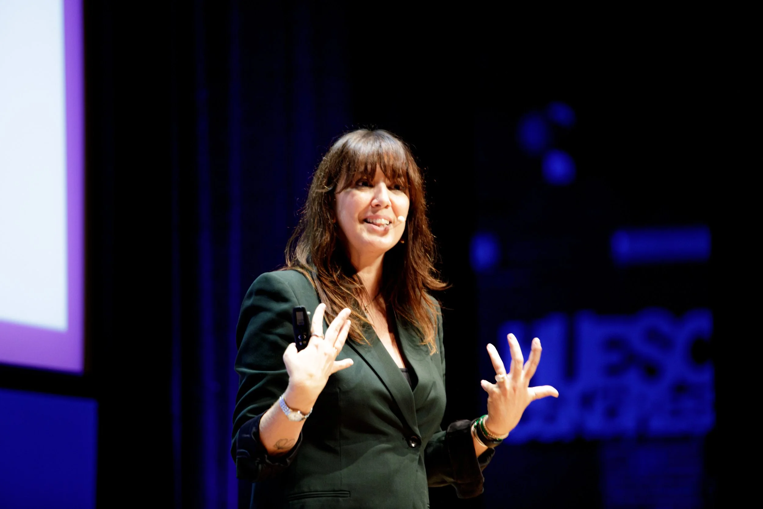 A woman with brown hair and bangs wearing a black blazer, speaking on stage with a headset microphone.