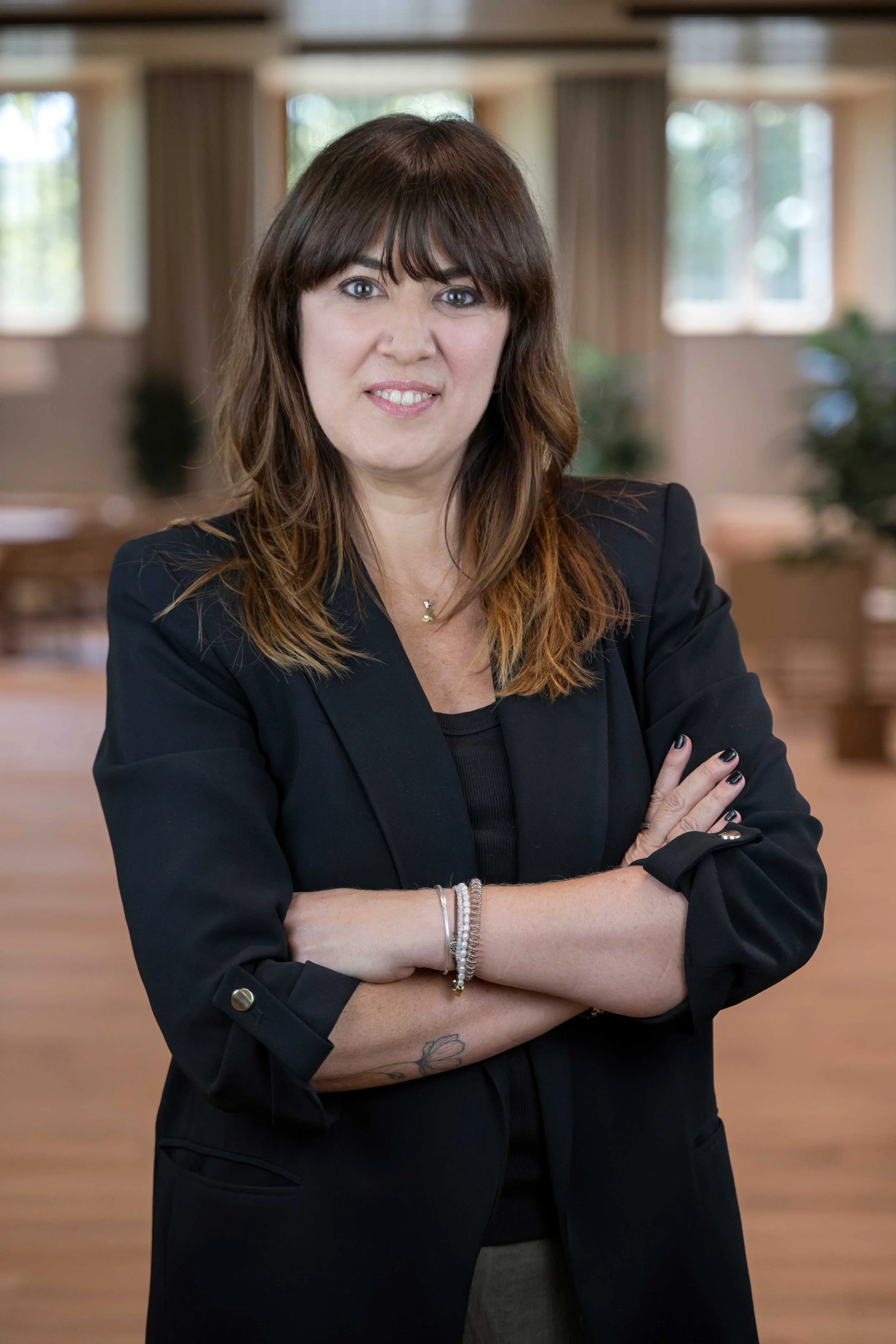 A woman with brown hair and bangs, wearing a black blazer, standing with arms crossed in a room with wooden floors and large windows.