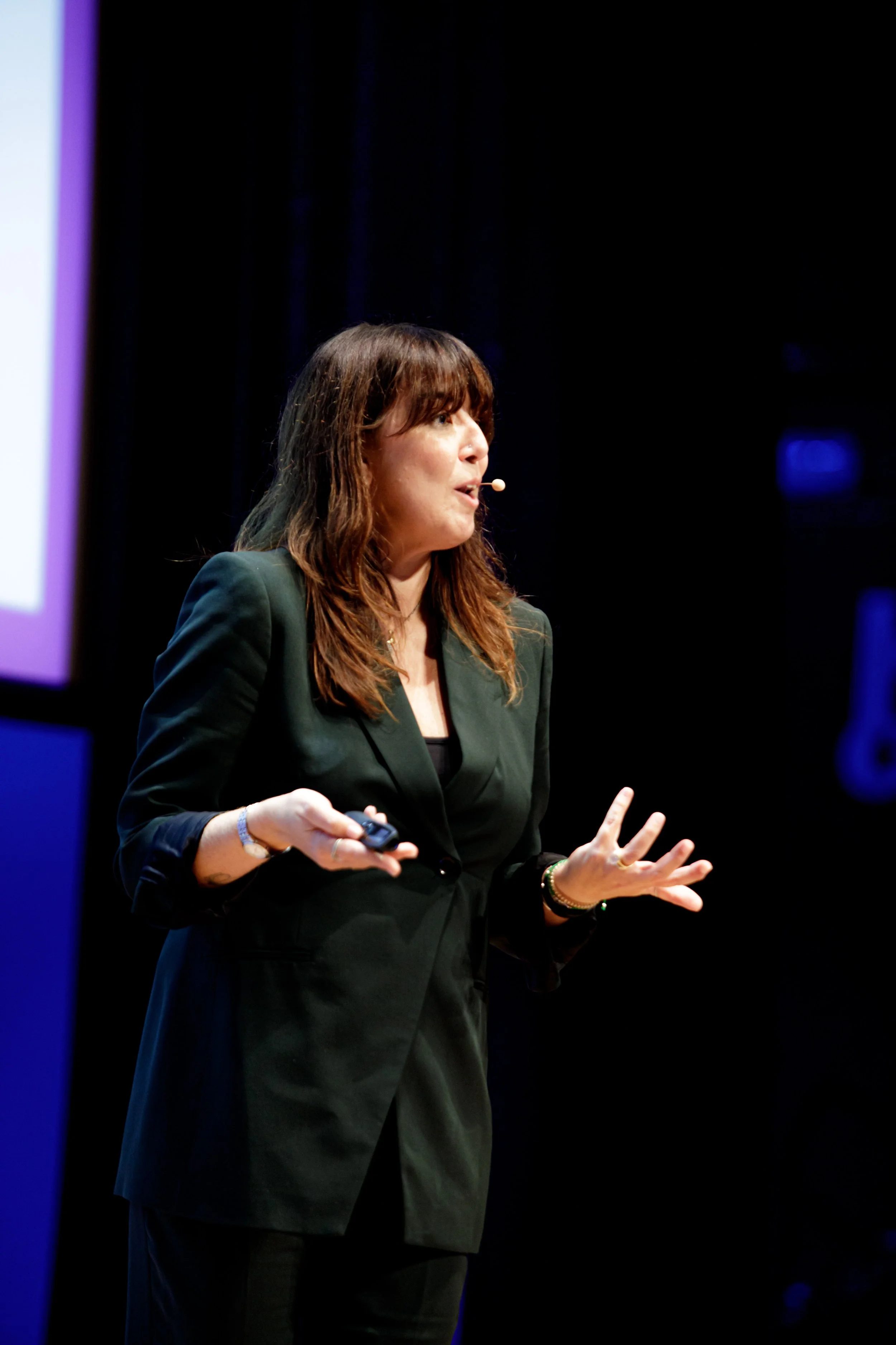 A woman with brown hair and a black blazer giving a presentation on stage, using a micro headset, with a dark background and a large screen to her side.
