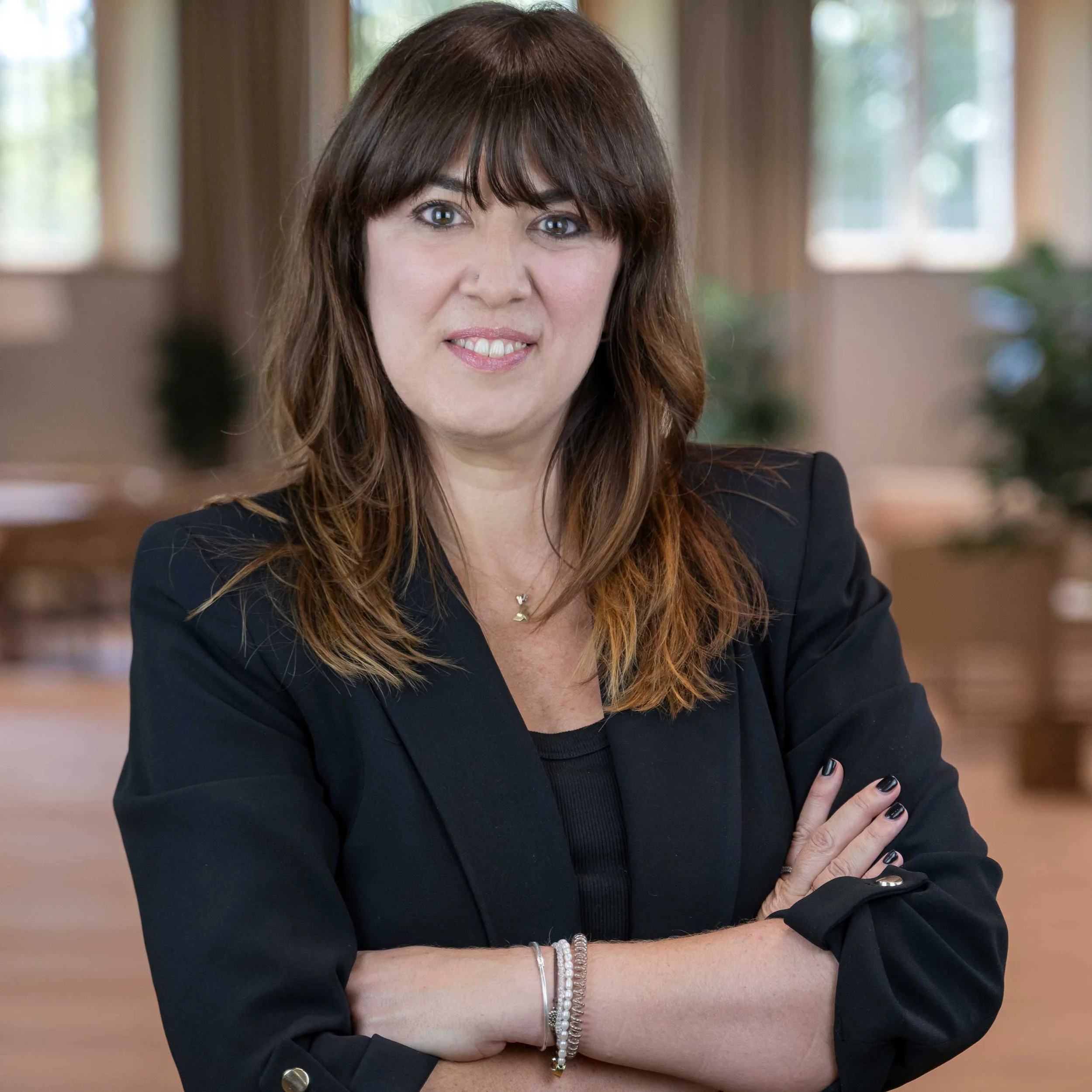 A woman with shoulder-length brown hair wearing a black blazer, standing with her arms crossed in a room with large windows and greenery outside.