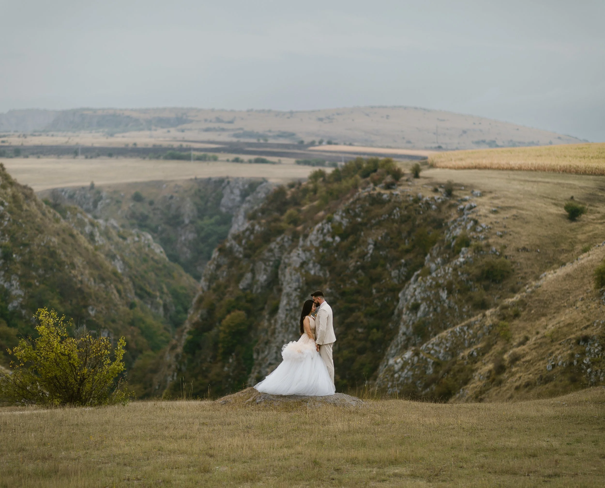 A couple dressed in wedding attire standing on a small rock in a scenic landscape with green hills and a canyon in the background.