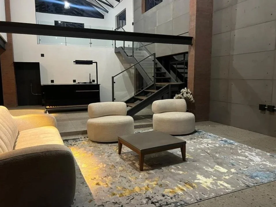 Modern indoor lounge area with beige sofa, two cream rounded chairs, a small coffee table, a patterned rug, and a staircase leading to an upper level with glass railings.